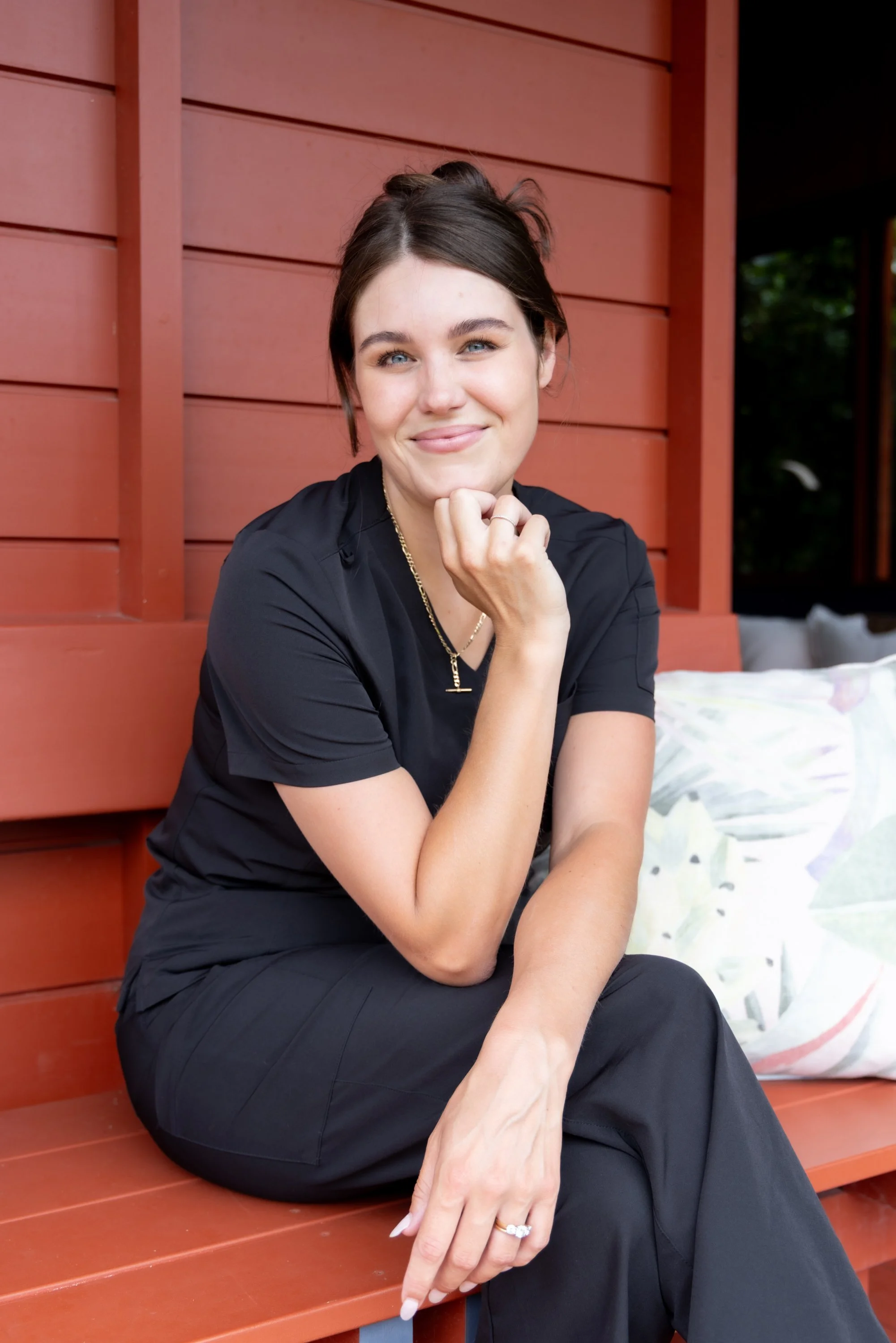 A woman with dark hair, wearing a black shirt and pants, sitting on a wooden bench with red slats, smiling, and looking at the camera. There is a cushioned pillow behind her and greenery visible in the background.
