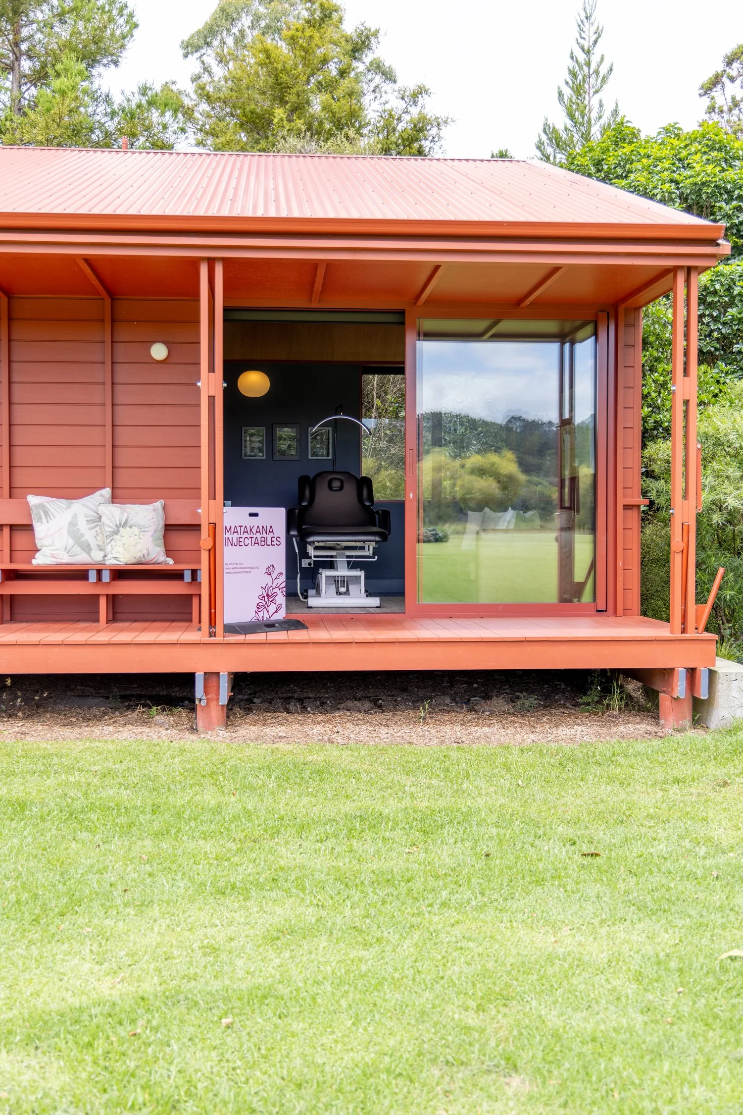 Small red house with a sliding glass door, porch with benches and pillows, lifestyle clinic with a chair inside, and a sign that says 'Matakana Injectables', set in a lush outdoor area with trees and grass.