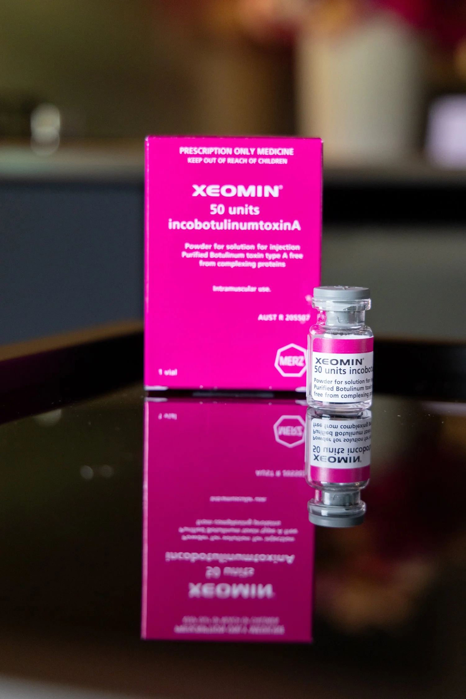 Vial of Xeomin medication and its pink box packaging behind it, both placed on a reflective surface.