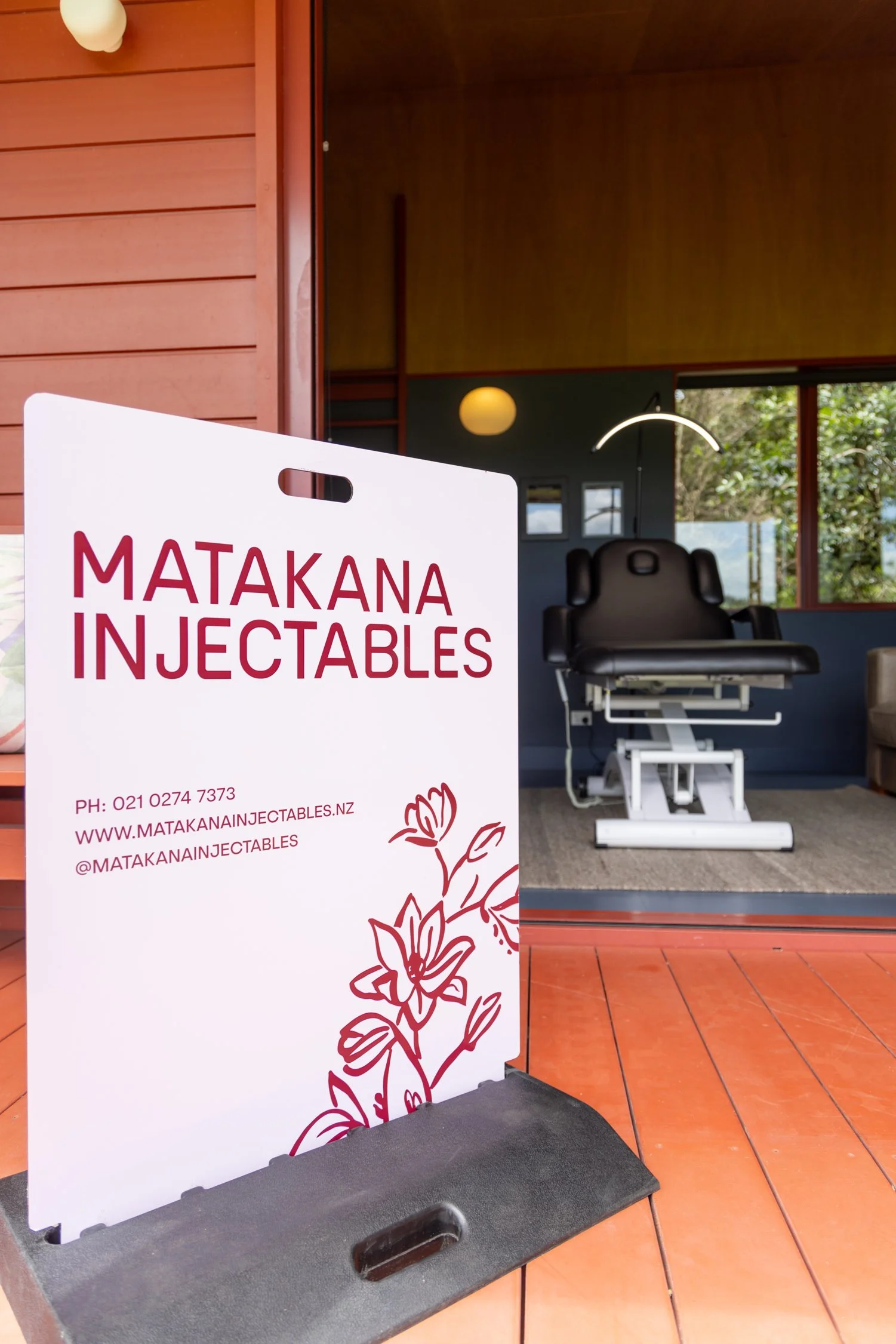 A signboard for Matakana Injectables with contact information and a floral design, placed outside a room with medical examination equipment visible inside.