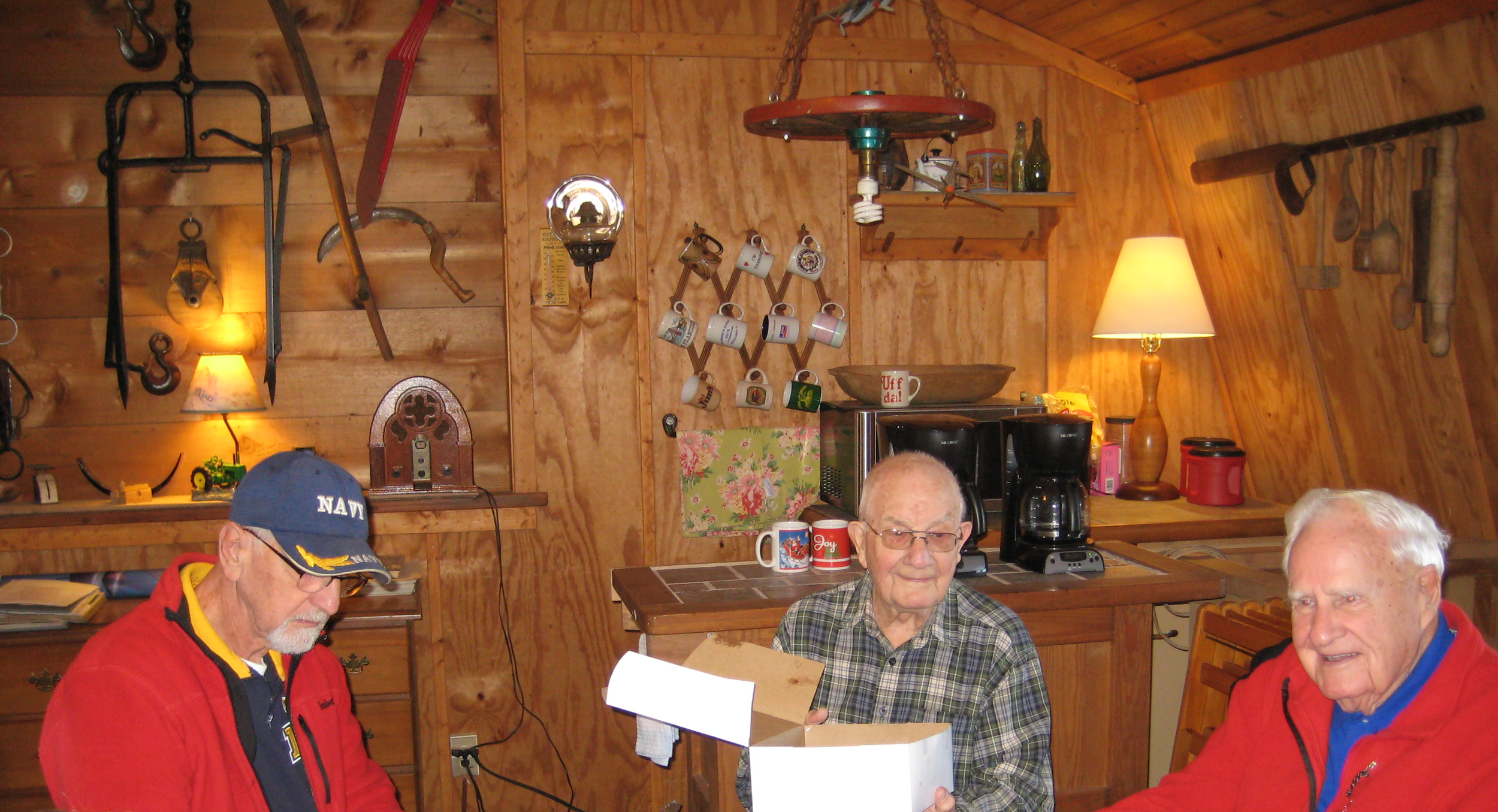 Three elderly men sitting in a cozy wooden barn, one holding a card and the others smiling. The kitchen has wooden walls, shelves with mugs, and vintage kitchen items including a radio, lamp, coffee maker, and various kitchen utensils.