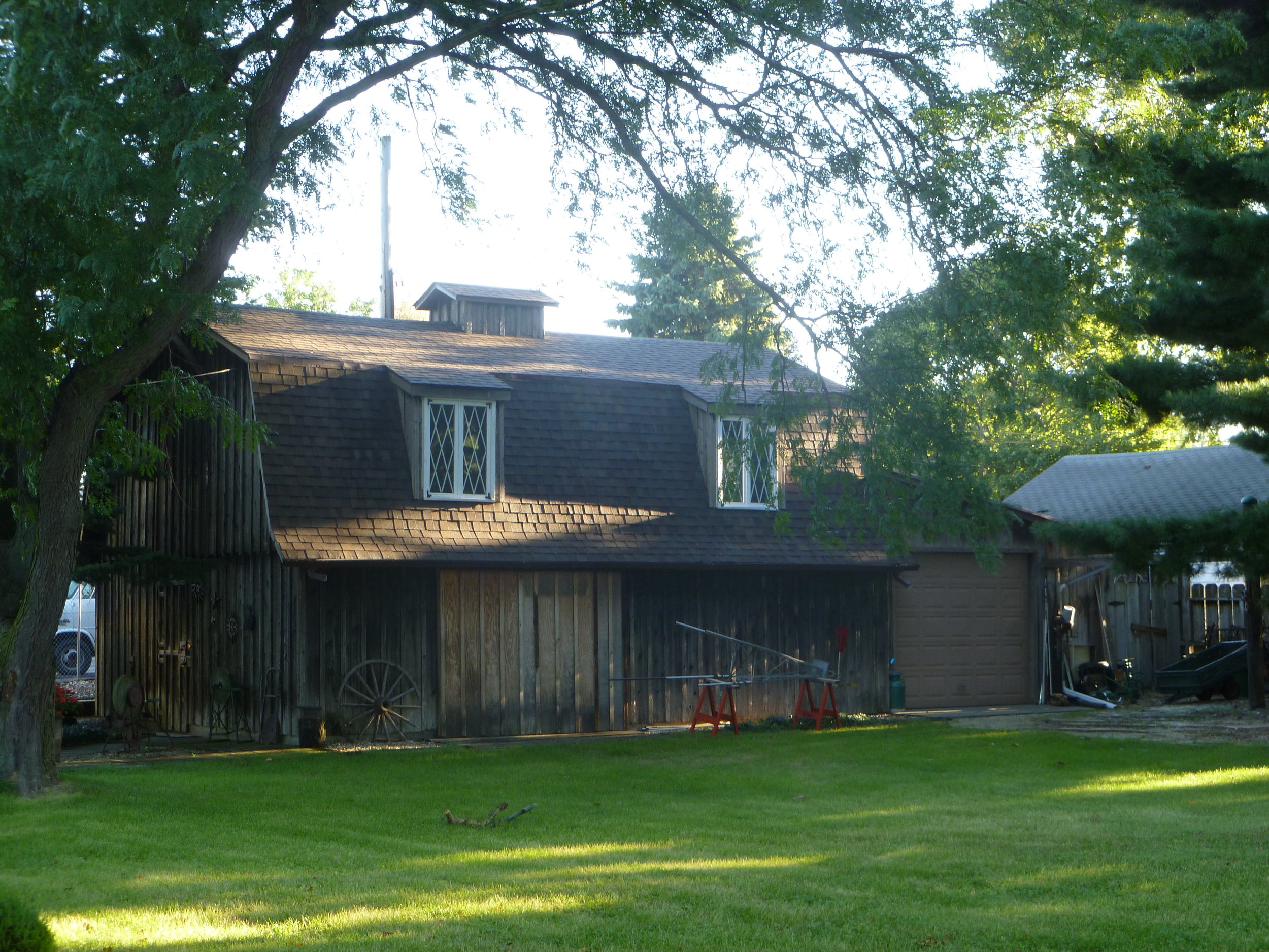 A rustic wooden barn with a shingled roof, two dormer windows, surrounded by large green trees and a well-kept lawn.