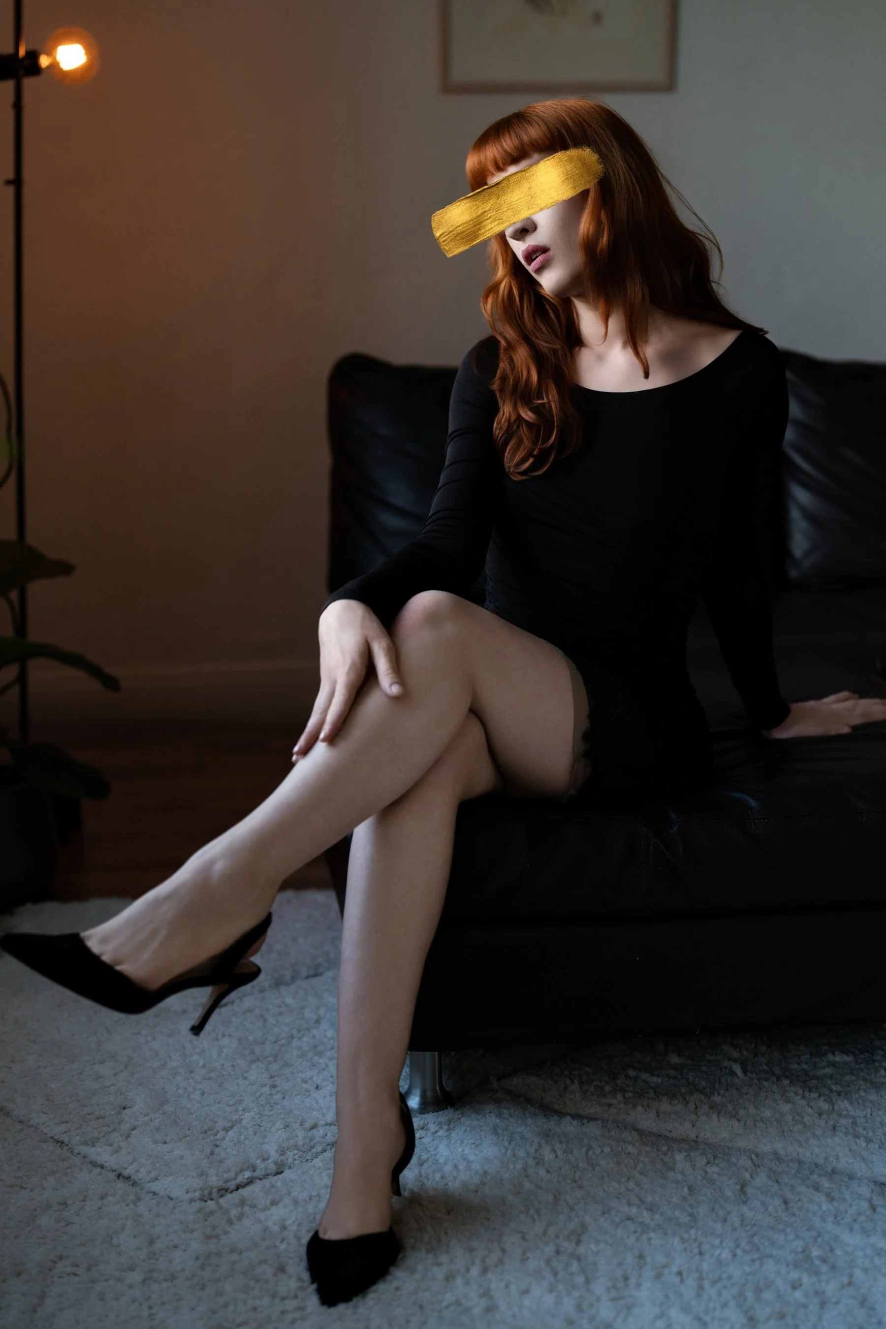 Claudia sits on a black leather couch, with red hair, wearing a short black dress. Her legs are crossed, trailing one hand up her top leg. The room is warmly lit, with a white rug undere the couch and art on the wall behind her.
