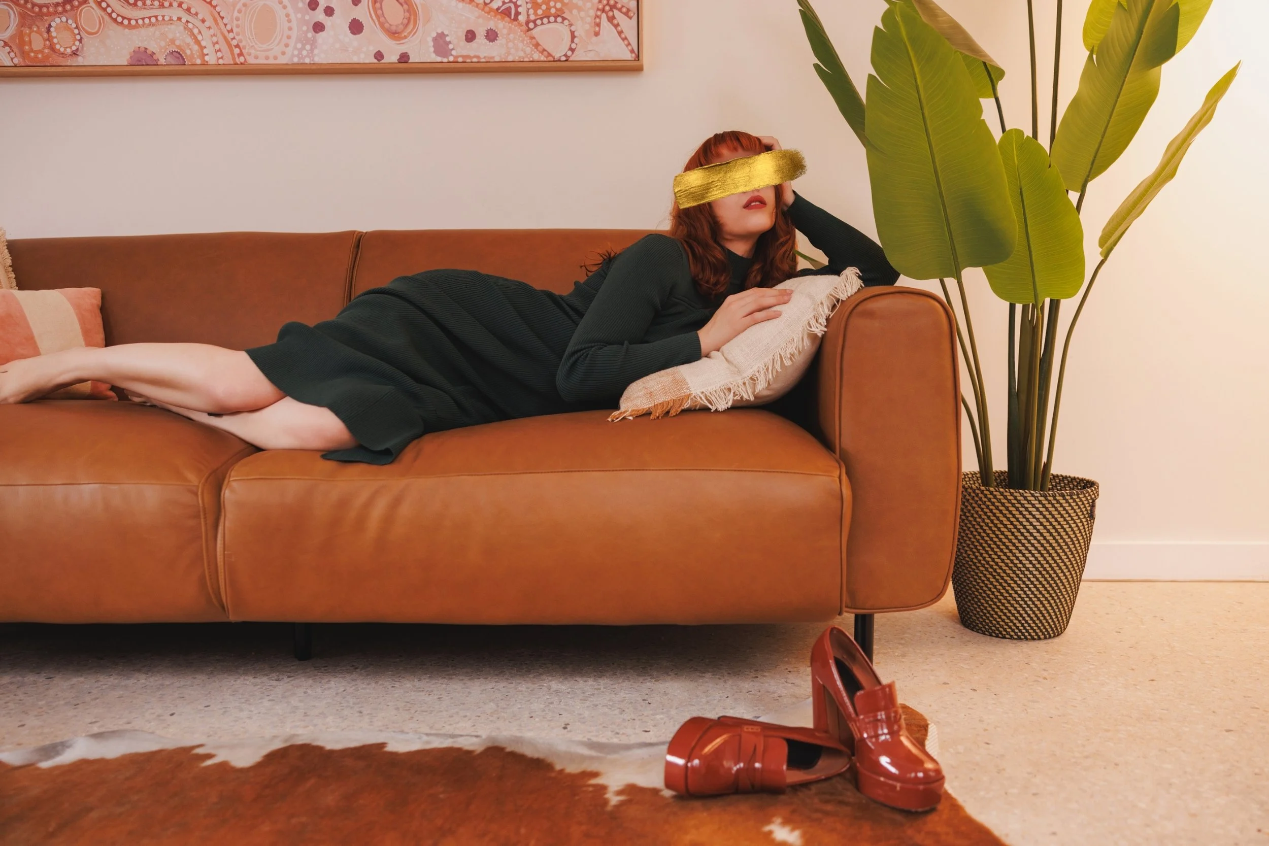 A woman with red hair, wearing a black dress, is lying on a brown leather couch with her eyes covered by a gold blindfold. She is resting on a pillow in a living room, with brown high-heeled shoes on the floor nearby, a large green plant beside the c