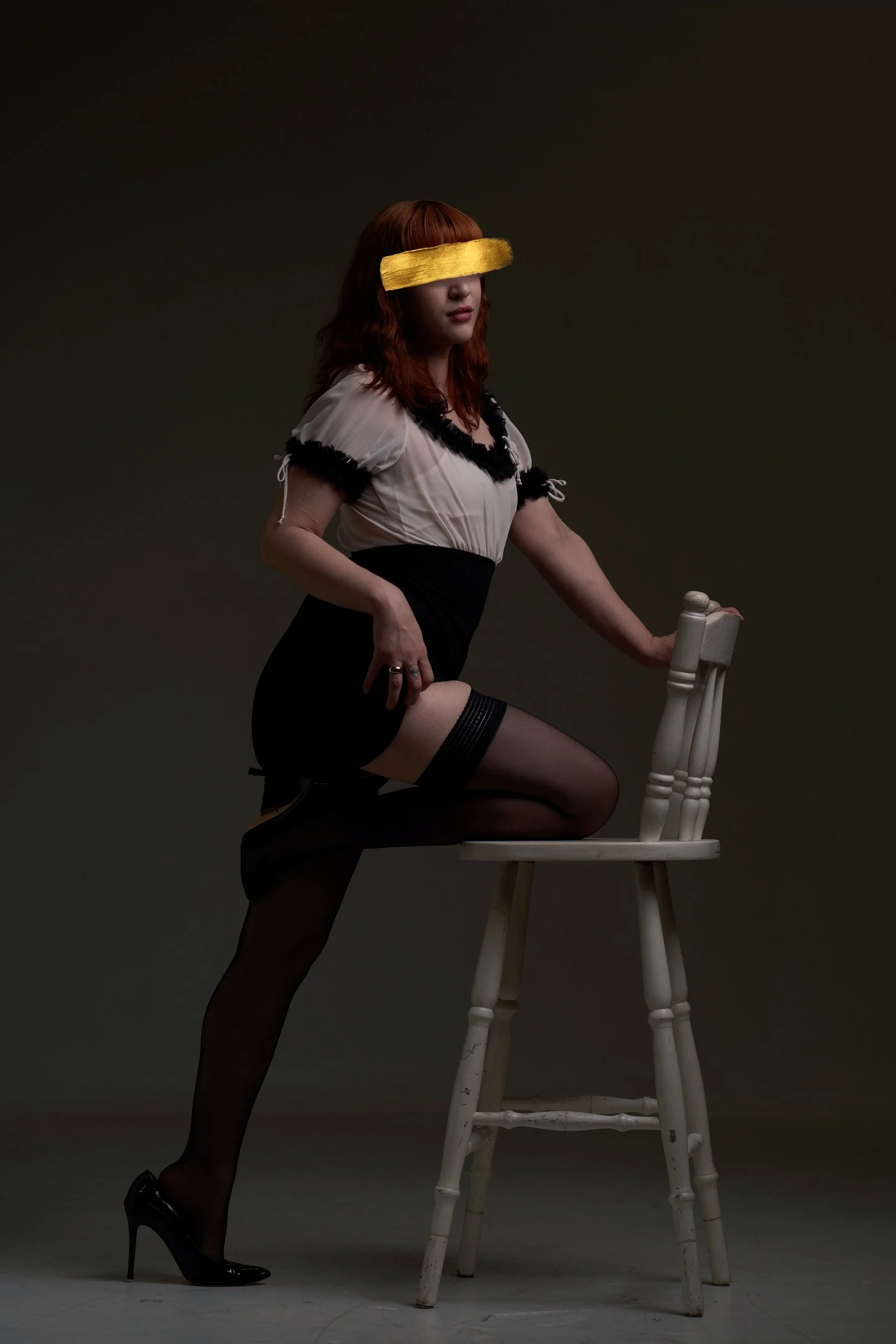A woman with red hair wearing black stockings, a black skirt, and a beige top with black trim, poses with one knee on a white wooden chair against a dark background, with a yellow blindfold covering her eyes.