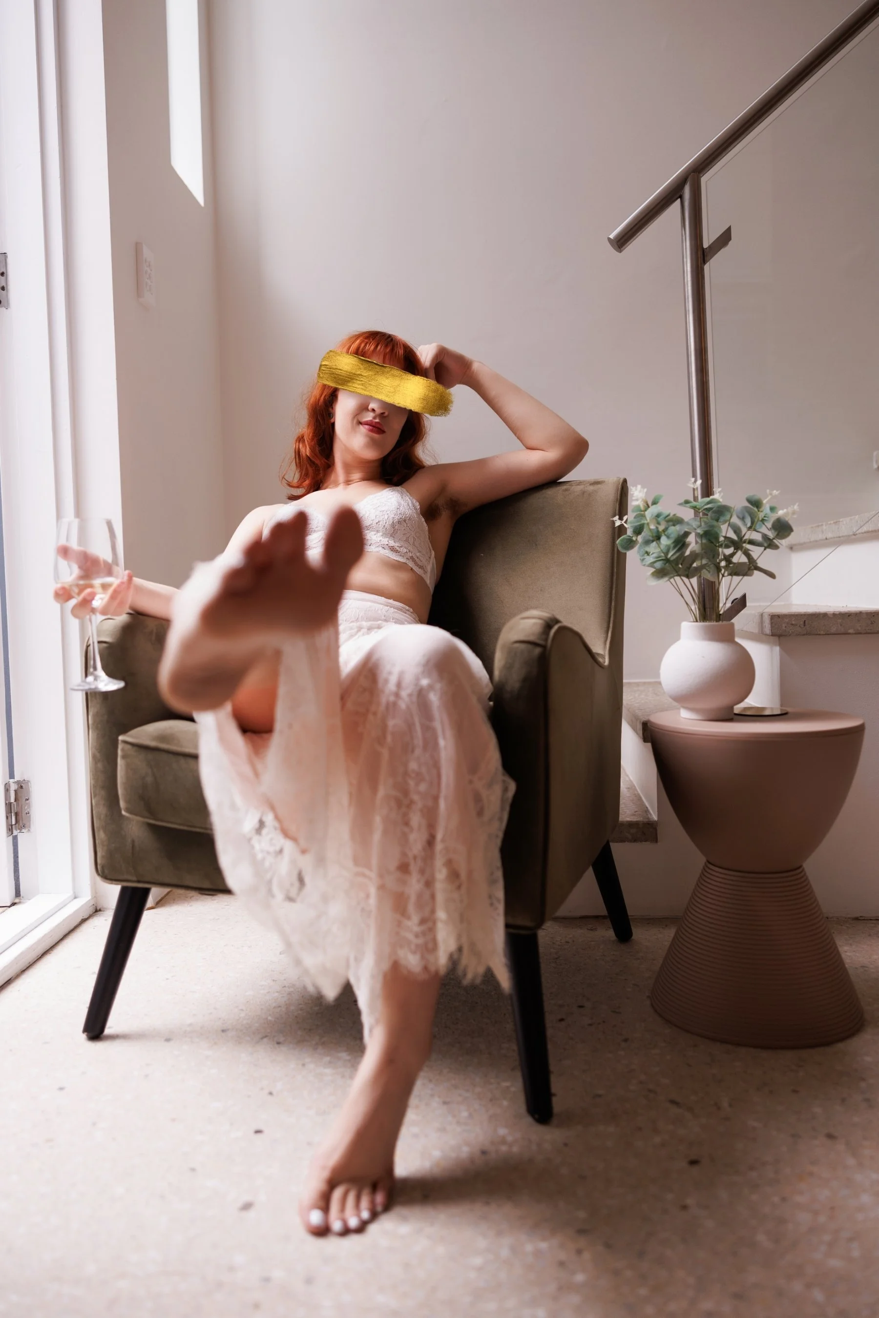 A woman with red hair, wearing white lace pajamas, sitting in an armchair beside a staircase with a potted plant on a small side table. Her eyes are covered with a yellow sleep mask, and she is holding a glass of wine with one foot resting on the arm
