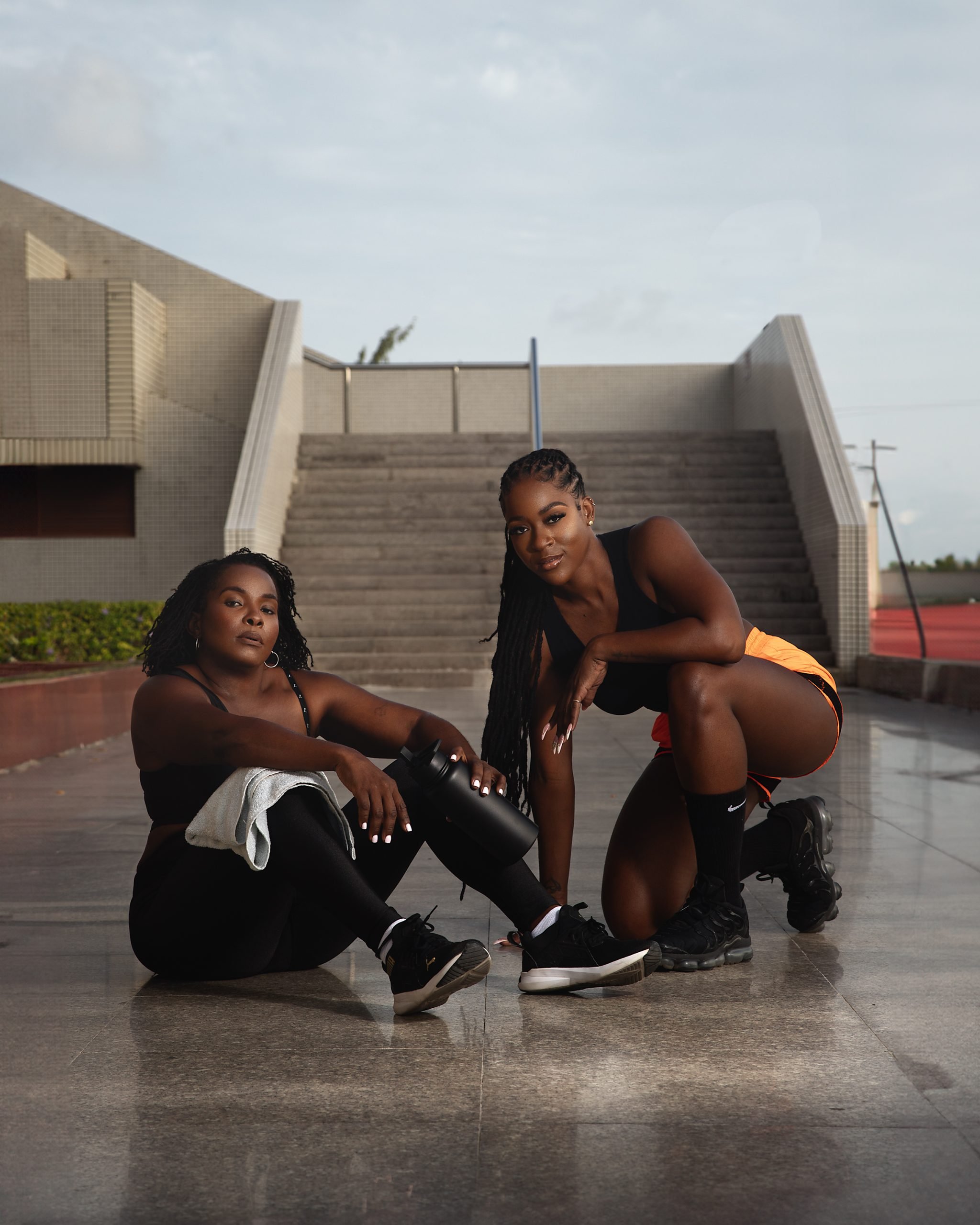 Two women dressed in athletic wear posing on a wet outdoor pavement near a staircase, with a modern building in the background.