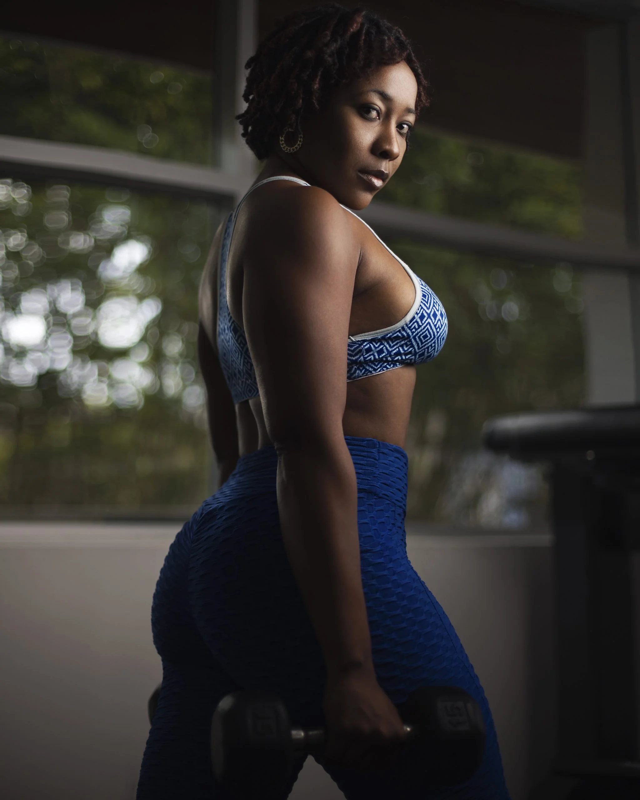 Woman in workout clothes holding a dumbbell in a gym with large windows and natural light.