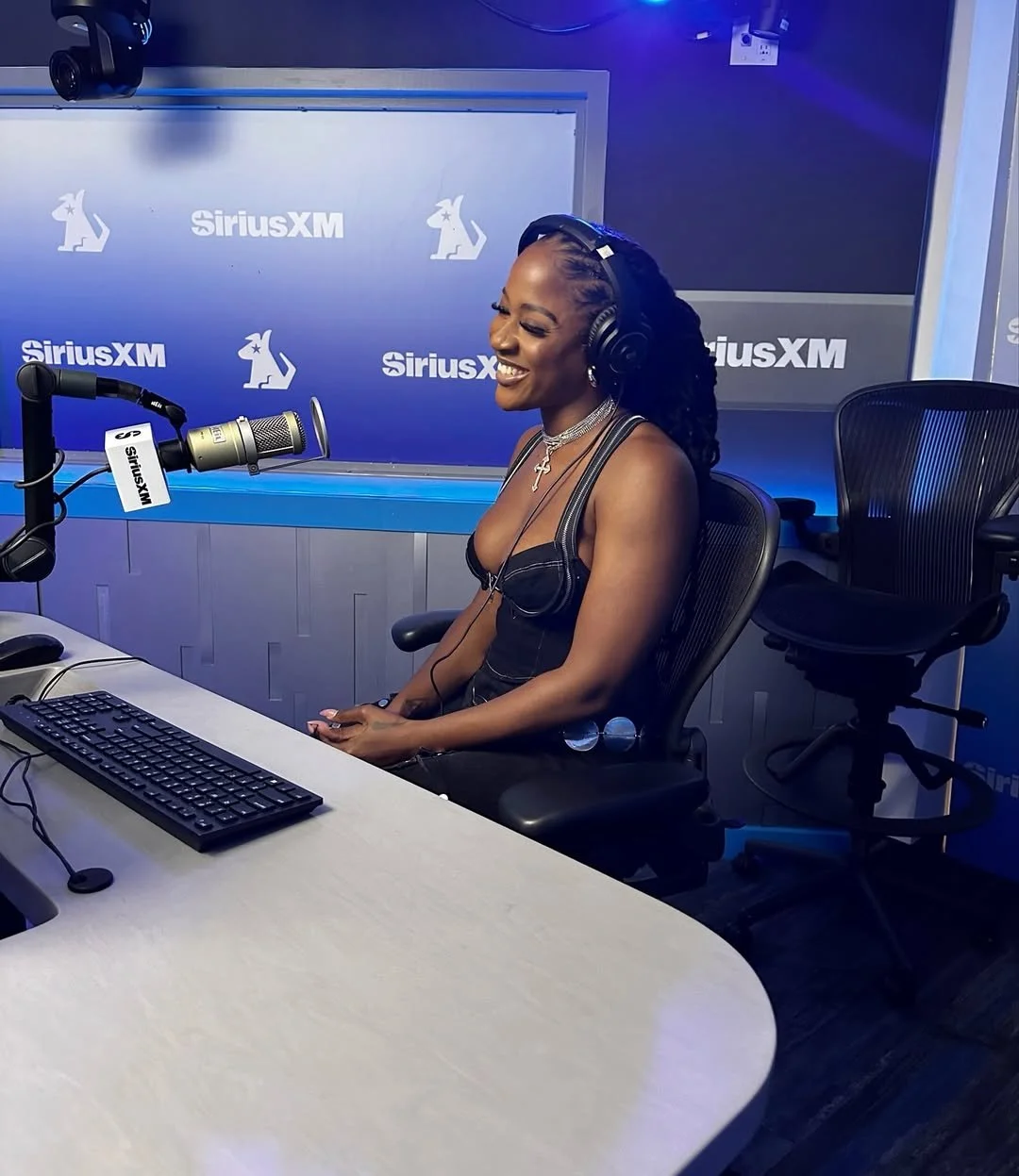 A woman with braids and jewelry is smiling and wearing headphones while sitting in a radio station studio with SiriusXM branding.