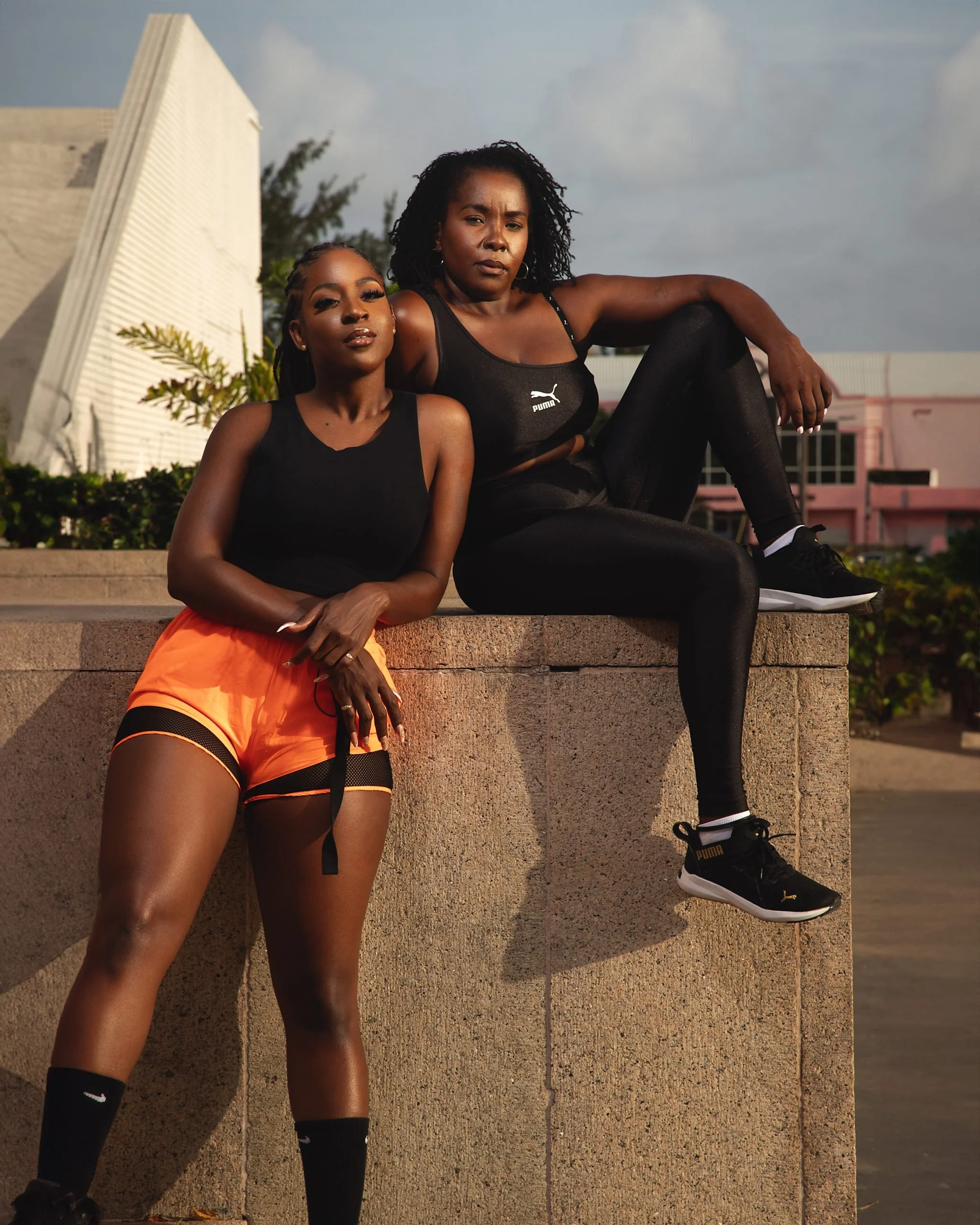 Two women in workout attire posing outdoors on a concrete ledge. One sits with legs bent, wearing black tank top and black leggings. The other stands with one leg propped on the ledge, wearing a black Puma top and orange shorts.