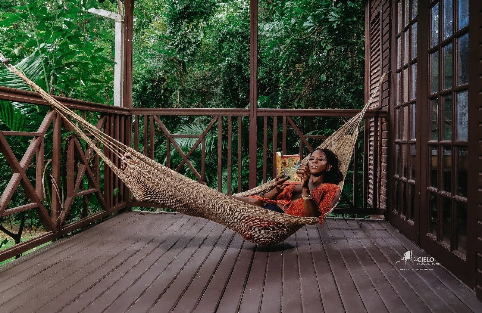 A woman relaxing in a hammock on a wooden balcony surrounded by lush green foliage, reading a book.