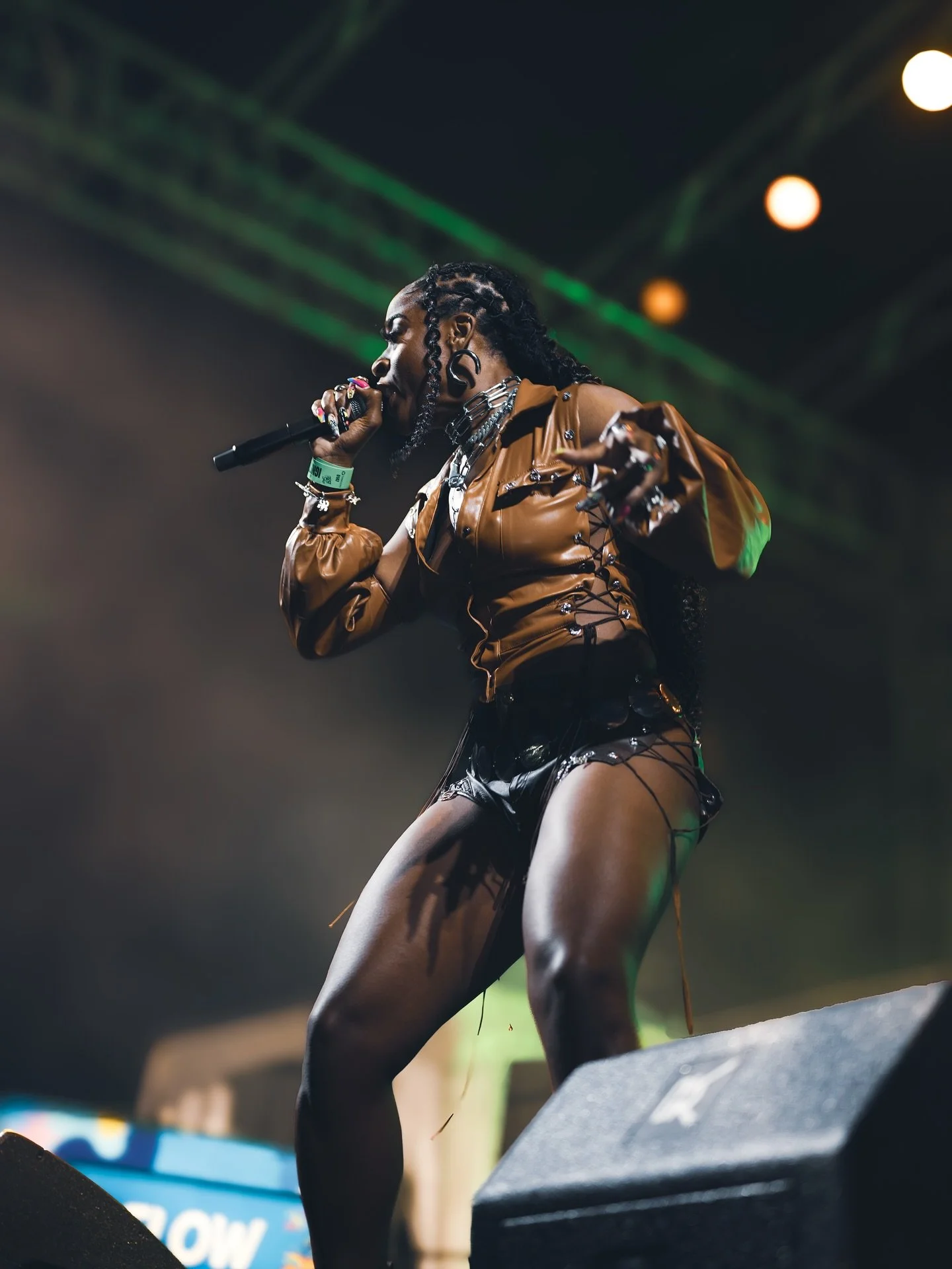 A female singer performing on stage with a microphone in hand, dressed in a brown leather jacket and black shorts, with braided hair and multiple jewelry accessories.