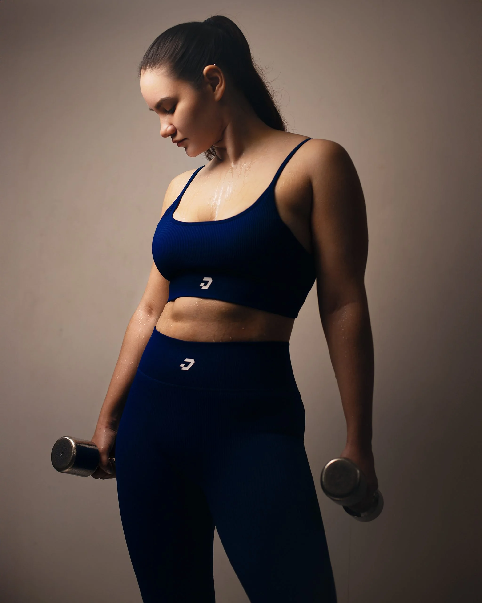A woman in black workout attire holding dumbbells, standing in a side profile pose, with sweat on her face and body against a plain background.