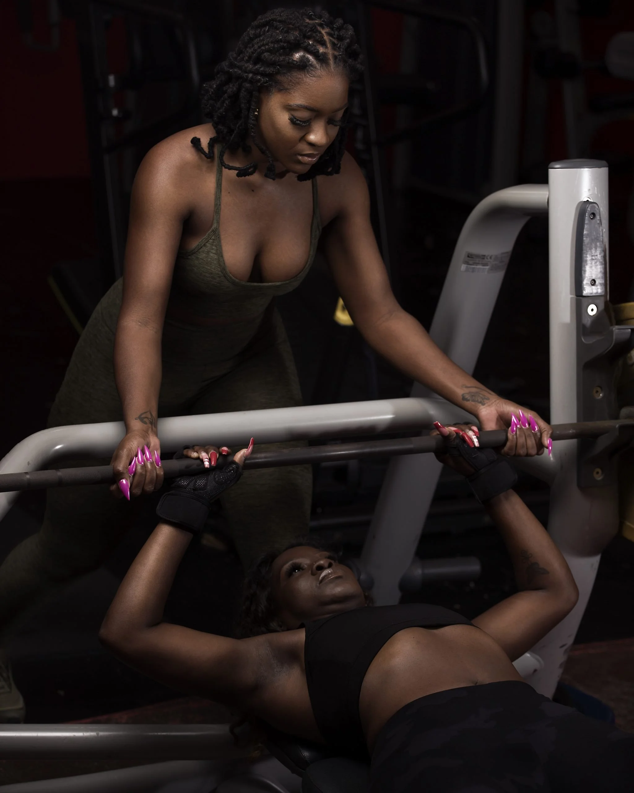 Women working out in a gym, one spotting the other during chest press exercise.