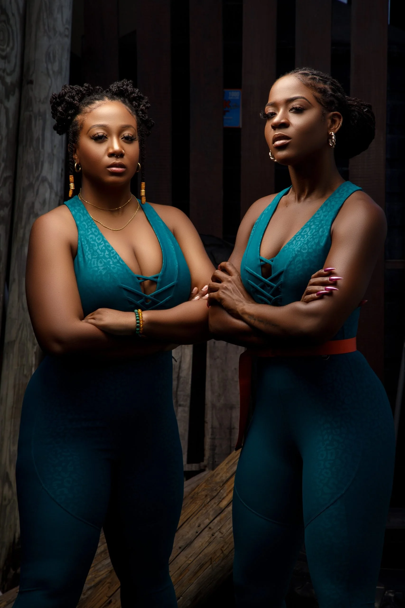Two women with dark skin, dressed in matching teal workout outfits, standing with crossed arms indoors against a dark wooden background.
