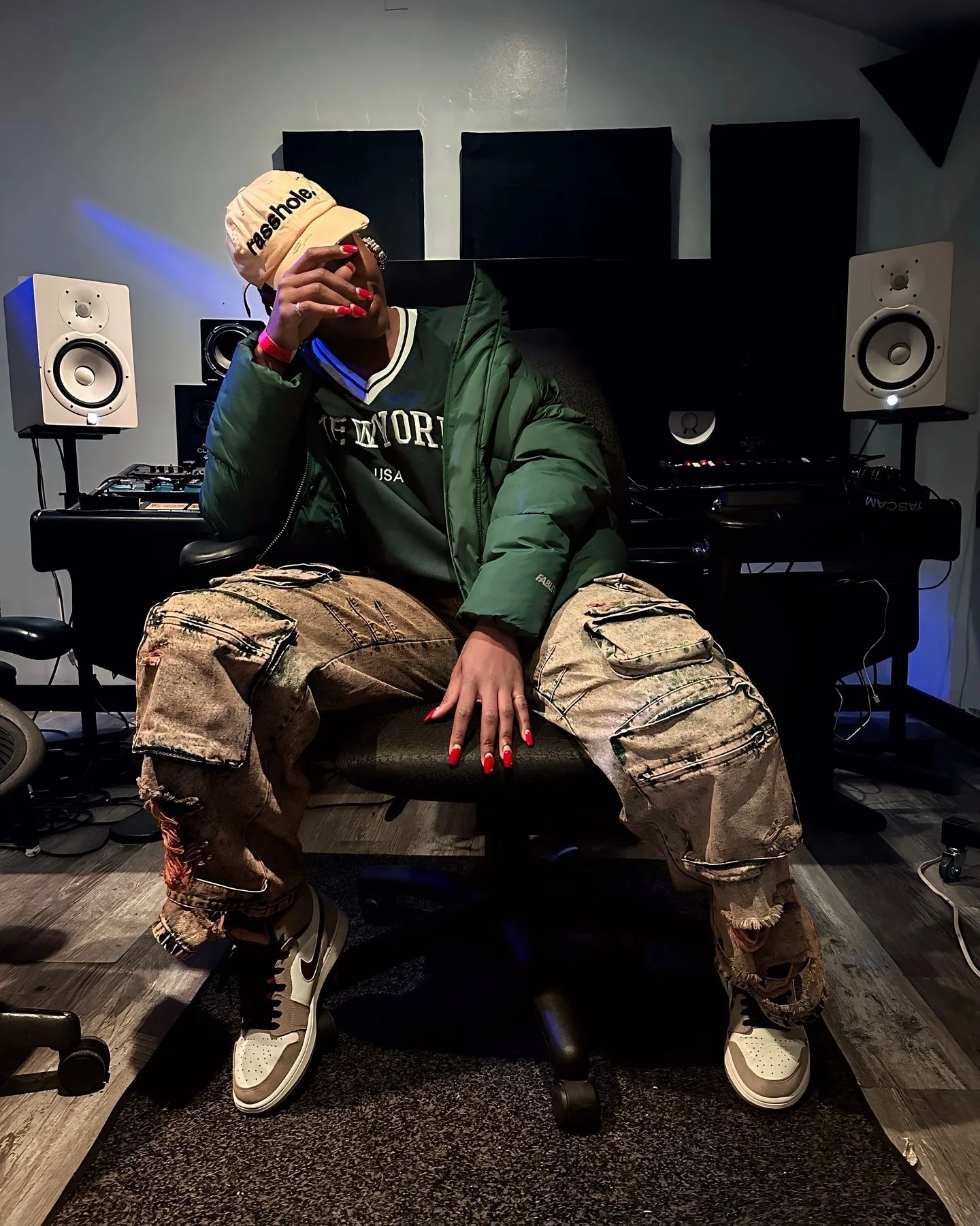 Person sitting on an office chair in a music studio, wearing a beige cap, green jacket, black sweatshirt, distressed cargo pants, and white sneakers, with hand covering face.