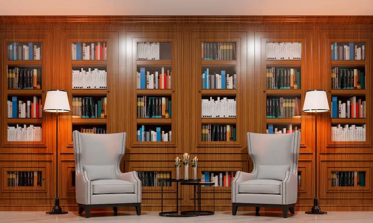A cozy library room with wooden bookshelves filled with books, two white armchairs, two floor lamps, and a small black table with a flower vase.