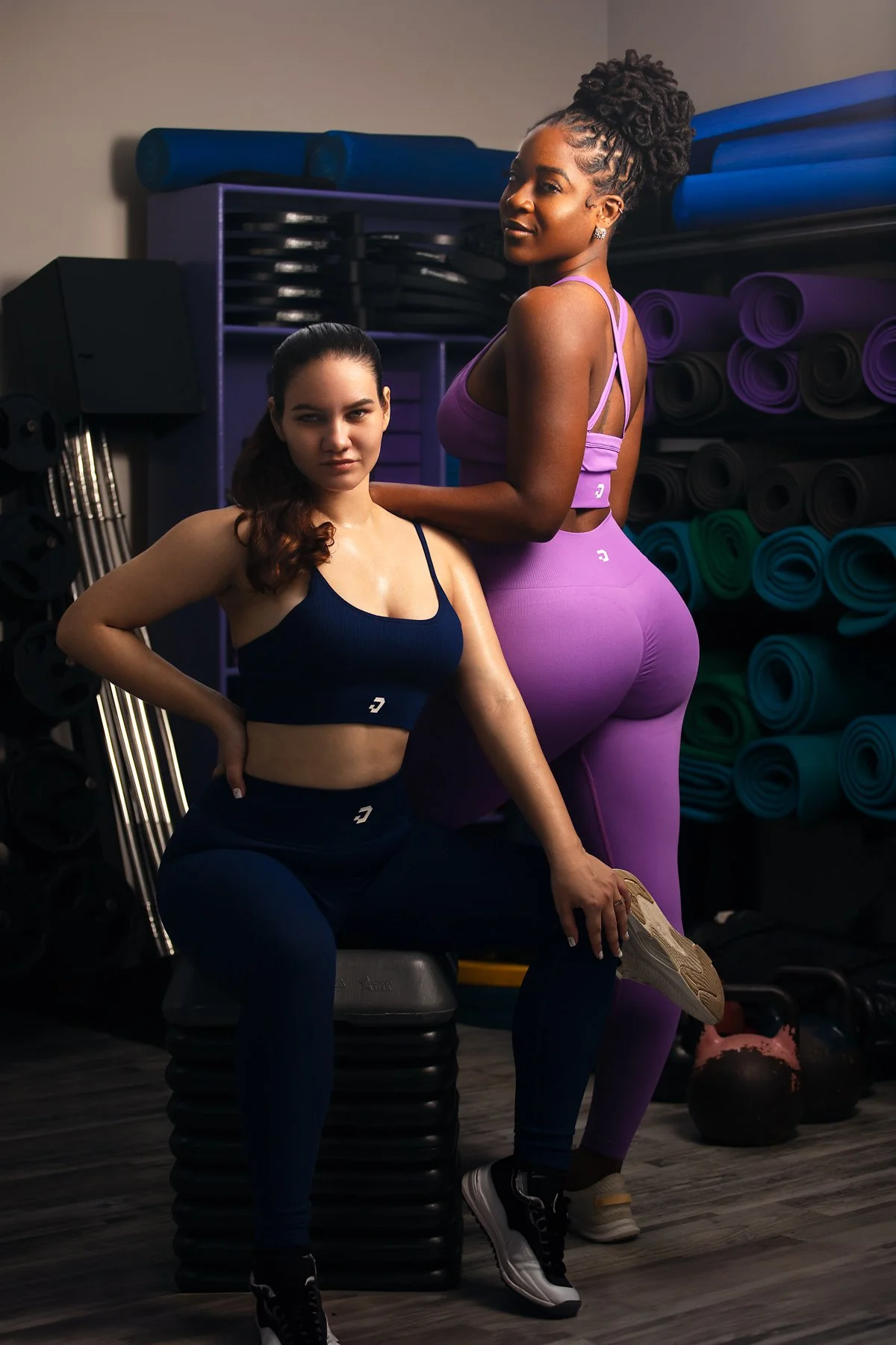 Two women in activewear in a gym, one seated on a black workout step, the other standing behind her. The seated woman is in navy blue sportswear, and the standing woman is in lavender workout clothing. Background has resistance mats and kettlebells.