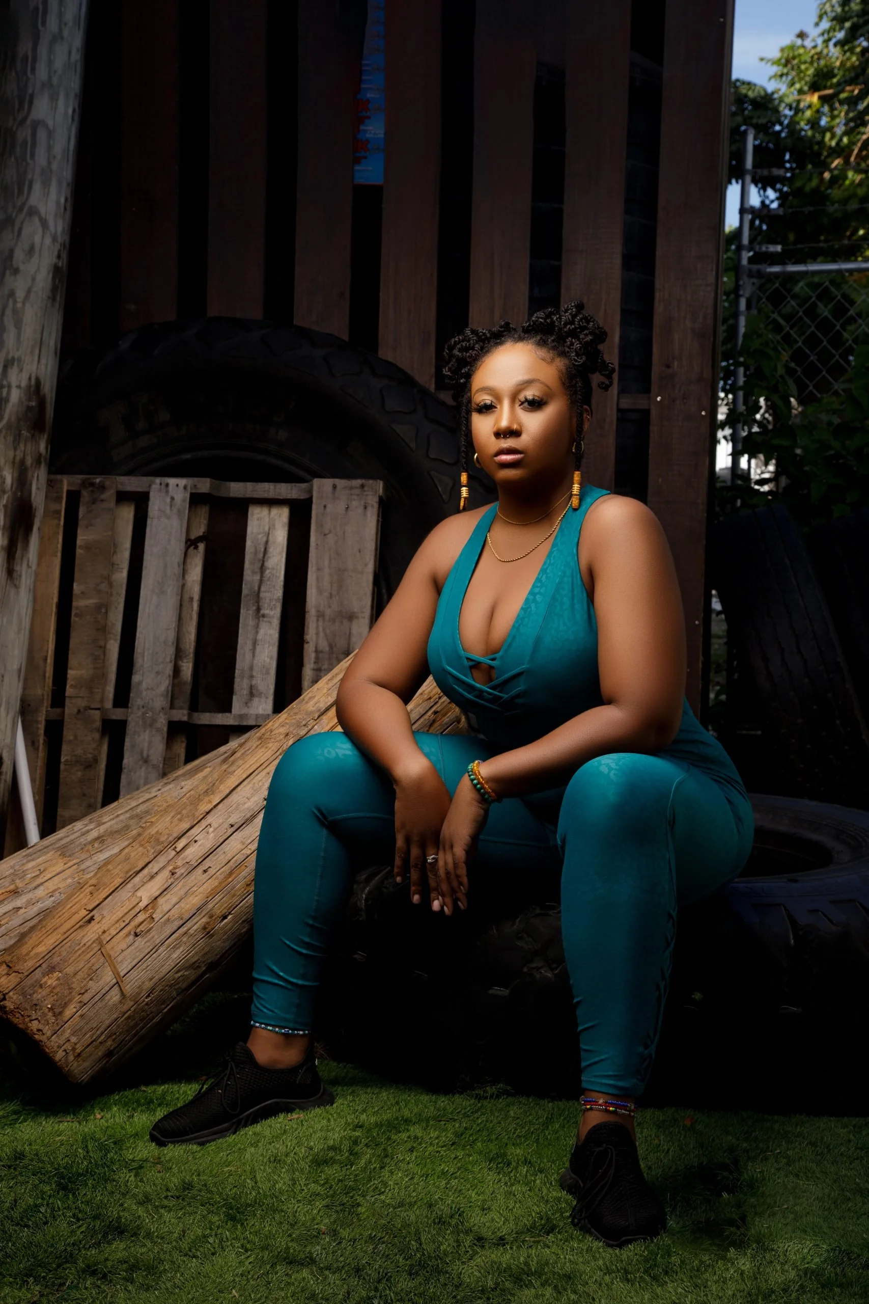 A young woman with braided hair, wearing a teal sleeveless jumpsuit, sitting outdoors on a wooden log with grass below. She has various bracelets and earrings, and her expression is serious as she looks at the camera. There are large tires and wooden
