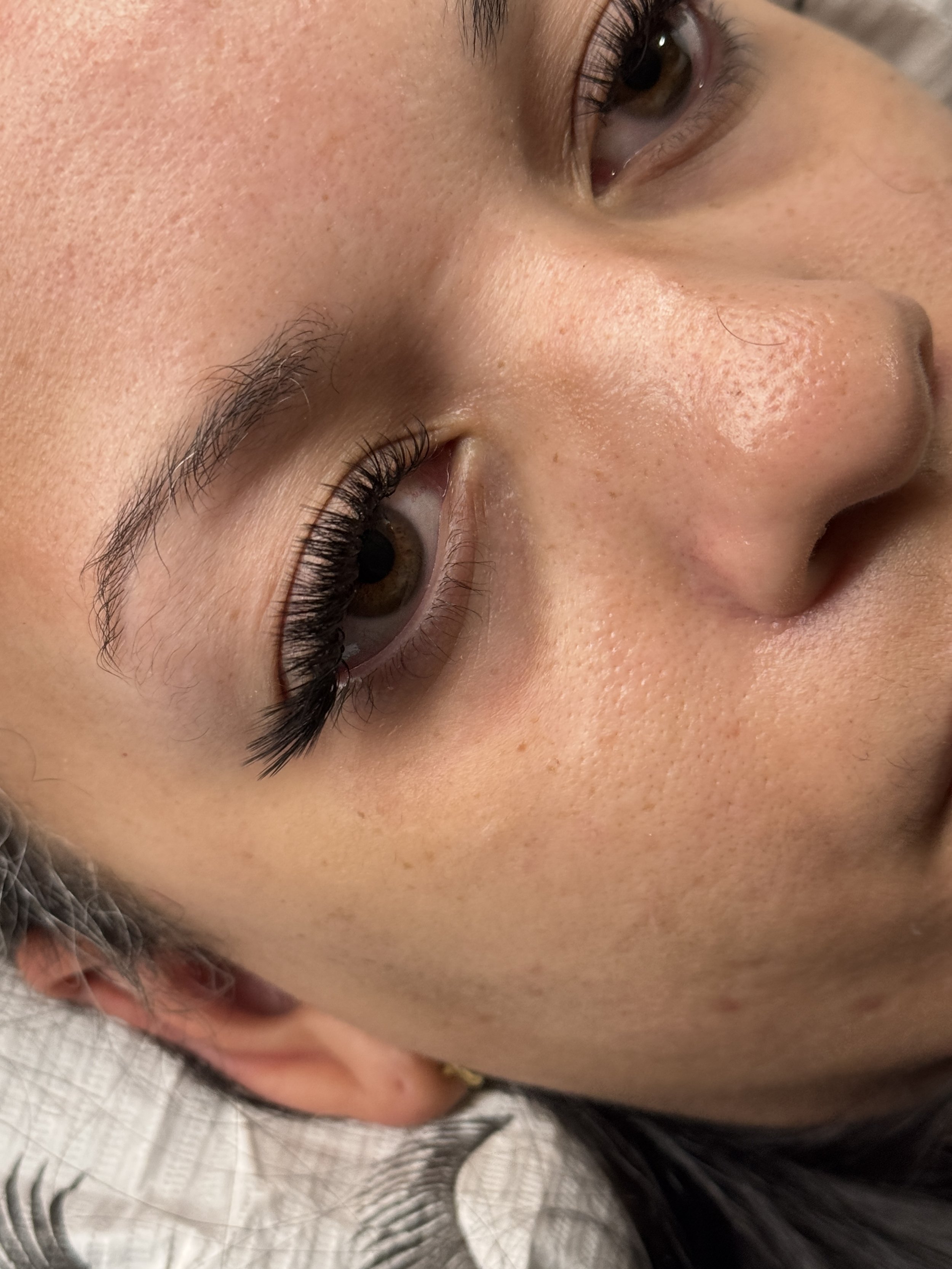Close-up of a person's face showing brown eyes, long eyelashes, smooth skin with visible pores, and part of an ear.