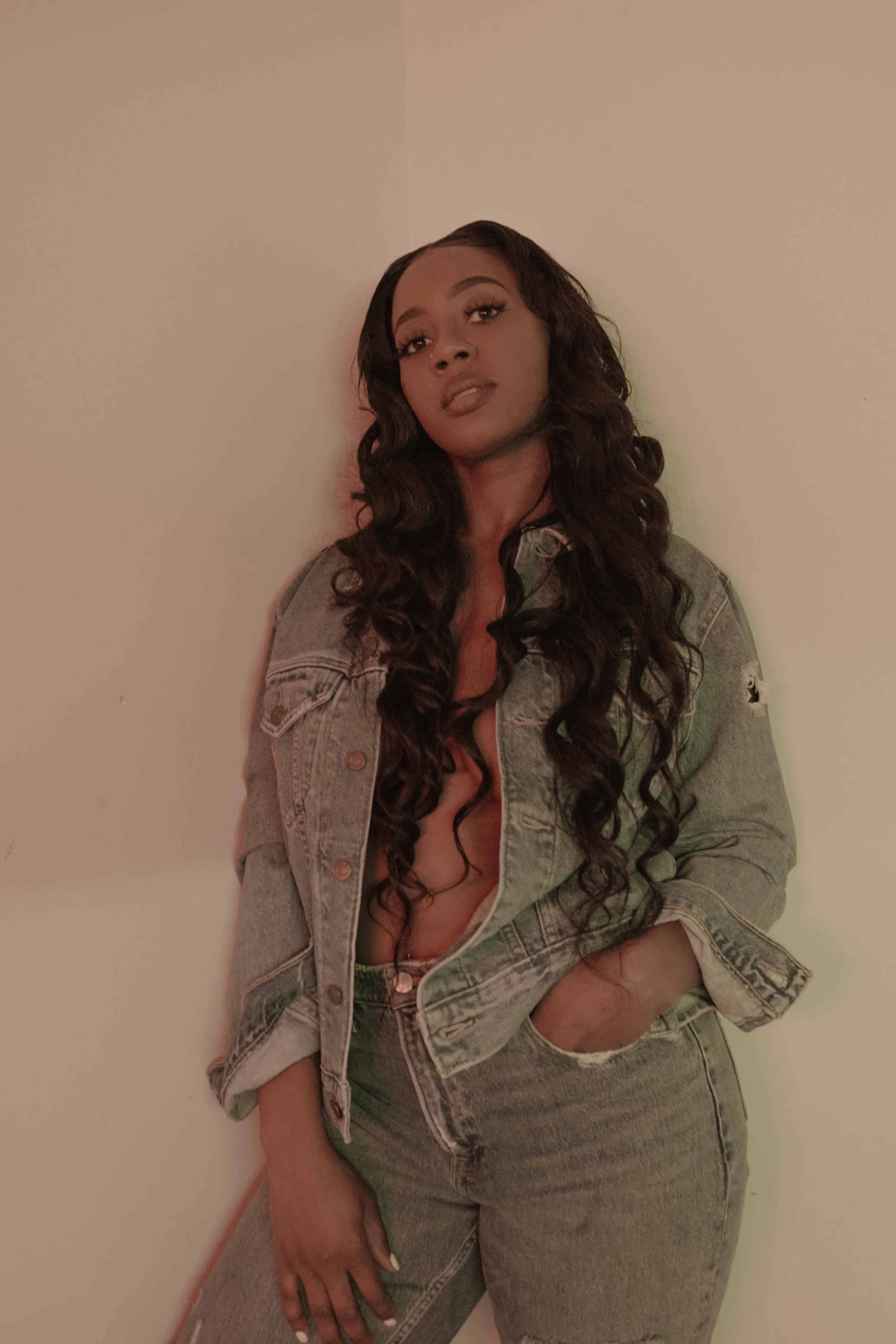 A woman with long curly hair wearing a denim jacket and jeans, standing against a plain wall.