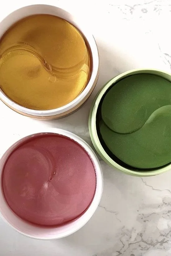 Three open containers of colorful hair styling gels or creams in yellow, green, and pink, placed on a white marble surface.