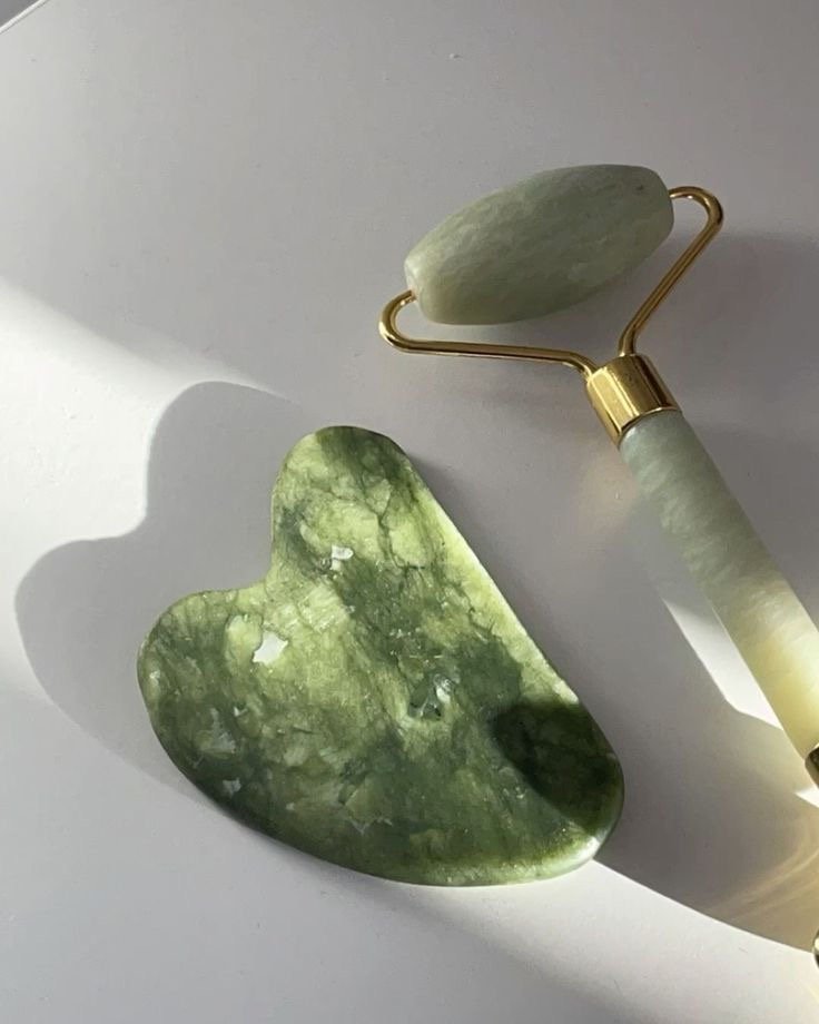 A jade roller and a jade gua sha tool on a white surface with shadows cast by natural light