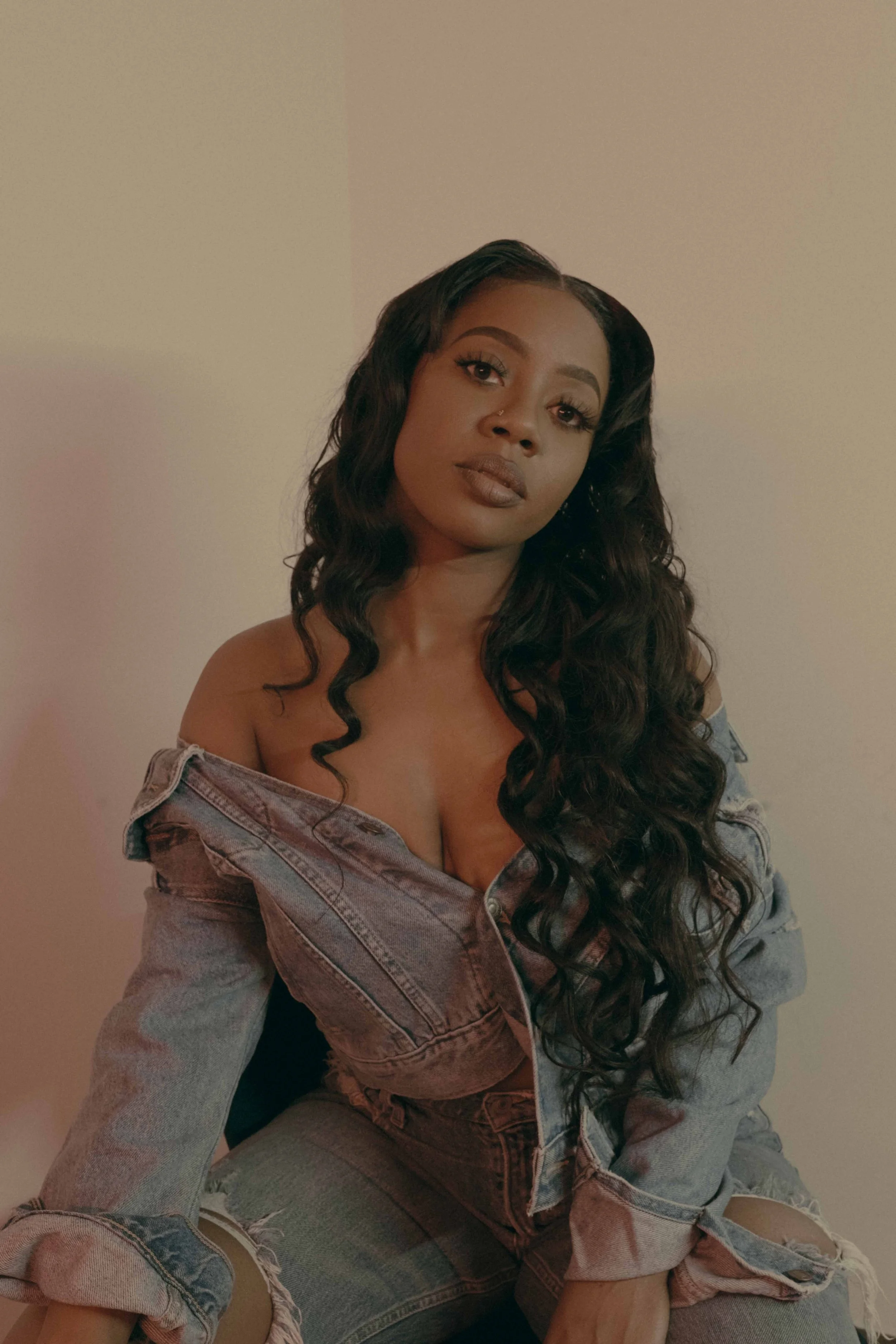A woman with long, curly hair wearing an oversized denim jacket and ripped jeans, sitting against a plain wall.
