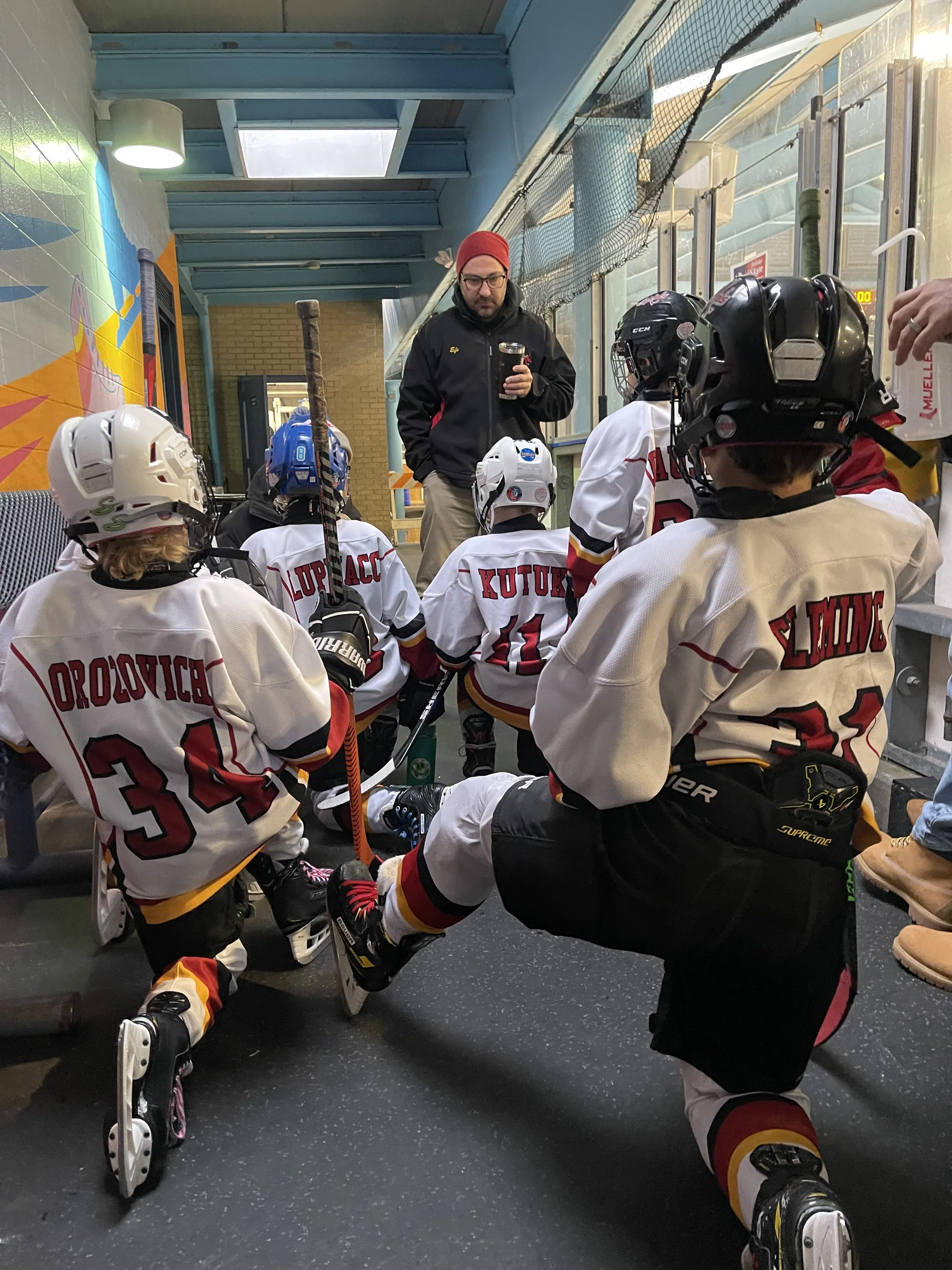 A youth hockey team gathered in the locker room, wearing white jerseys with red and black details, sitting on the floor, and listening to their coach who is standing and holding a cup, getting ready for or during a game.