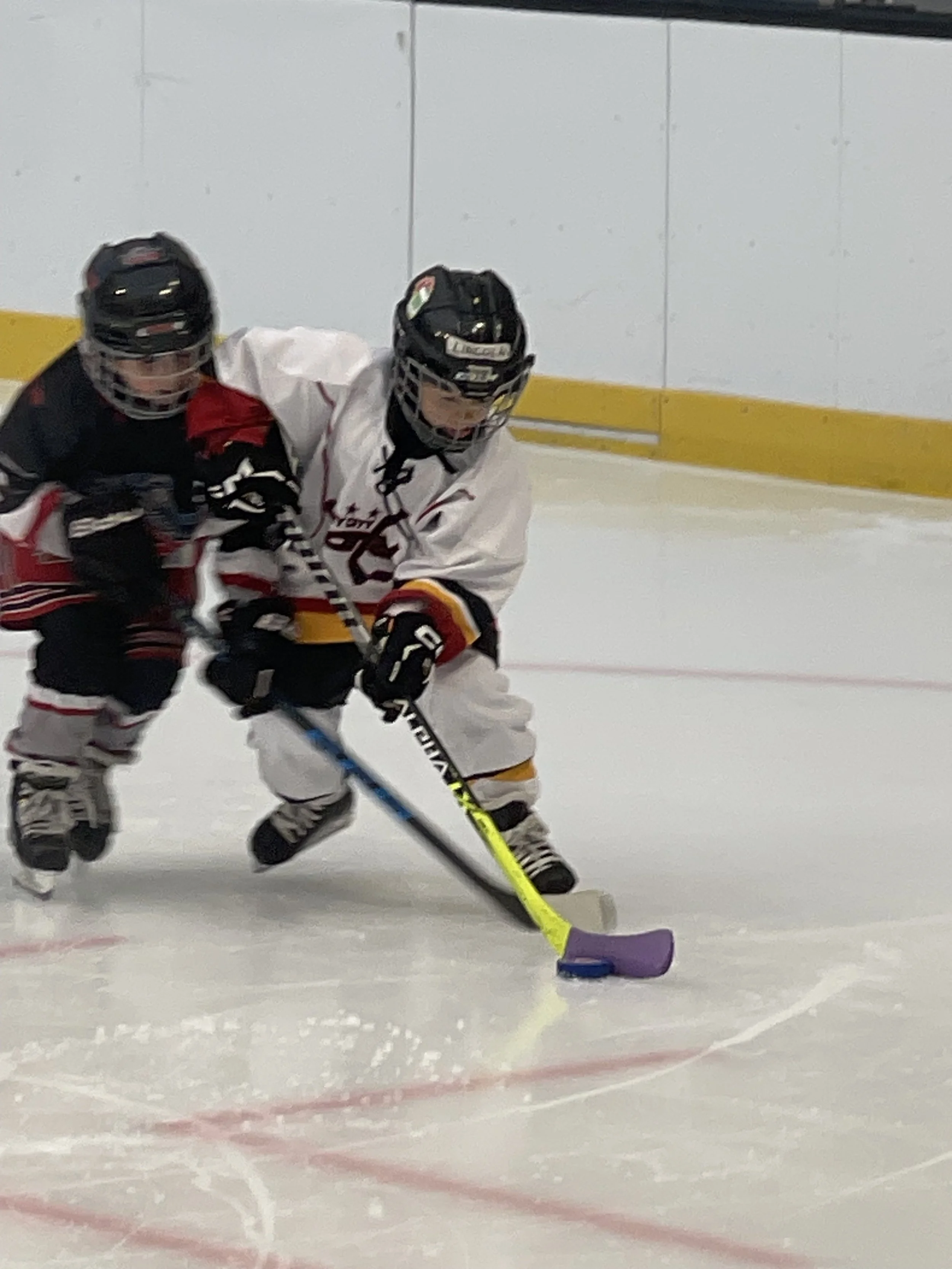 Two young hockey players competing for puck on ice rink, one in black and red uniform and the other in white and yellow uniform, both wearing helmets and skates.
