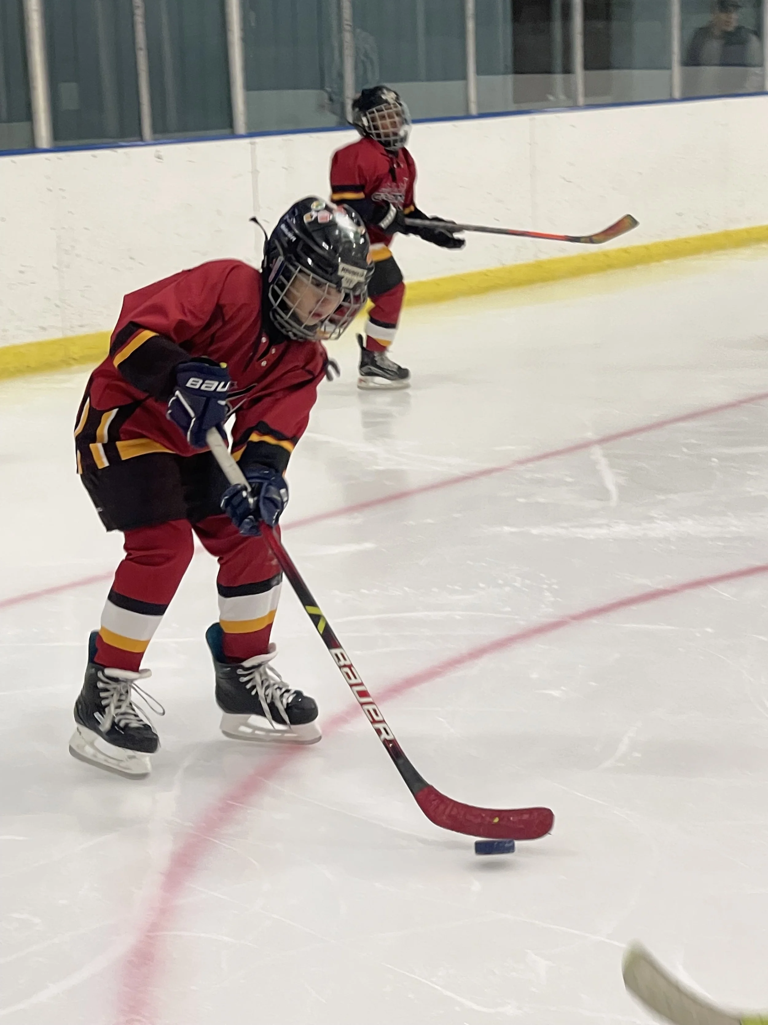Two young ice hockey players in red uniforms and helmets on an ice rink, one in the foreground controlling the puck with a hockey stick, and one in the background skating.