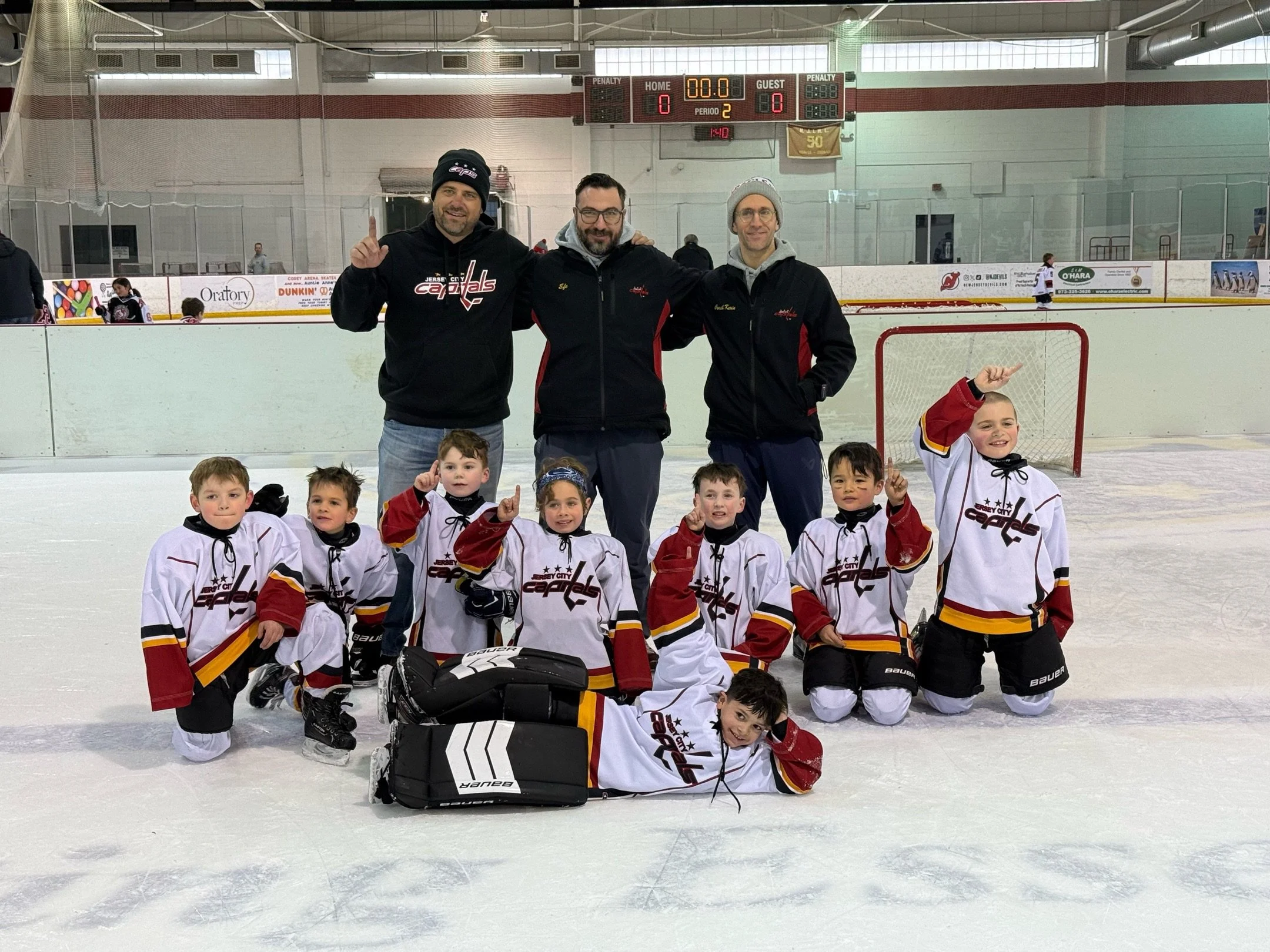 A youth hockey team with their coaches on an ice rink, posing after a game. The team members wear white jerseys with red and black details, kneeling and lying on the ice, smiling and making celebratory gestures. The coaches stand behind them, also smiling and dressed in dark jackets and hats. The scoreboard in the background shows a tied game.