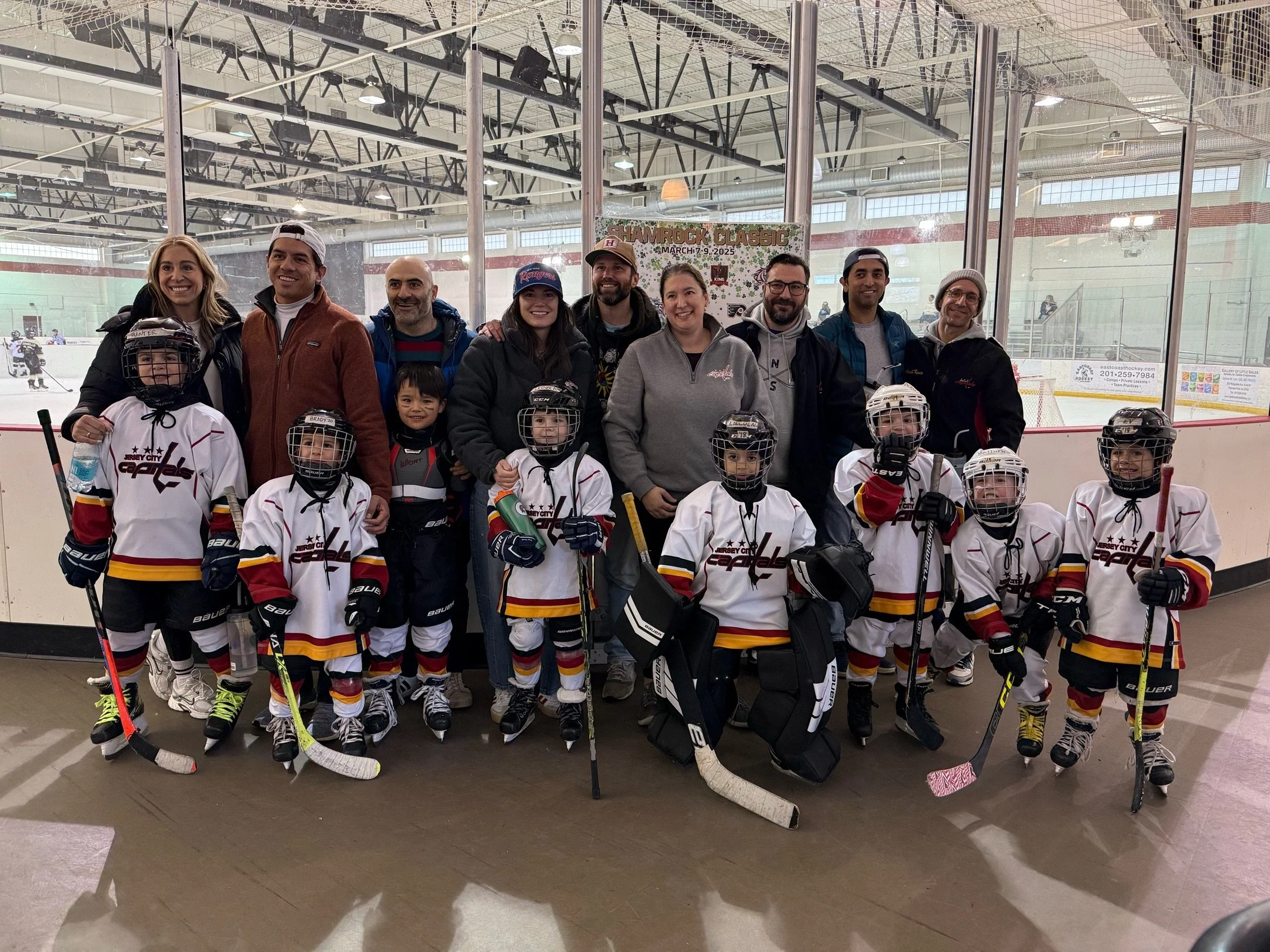 A group of young hockey players and their coaches posing together inside an ice rink. The children are wearing hockey jerseys, helmets, and skates, holding hockey sticks. The coaches and adults are standing behind them, smiling. In the background, there are other players on the ice and an ice hockey rink.