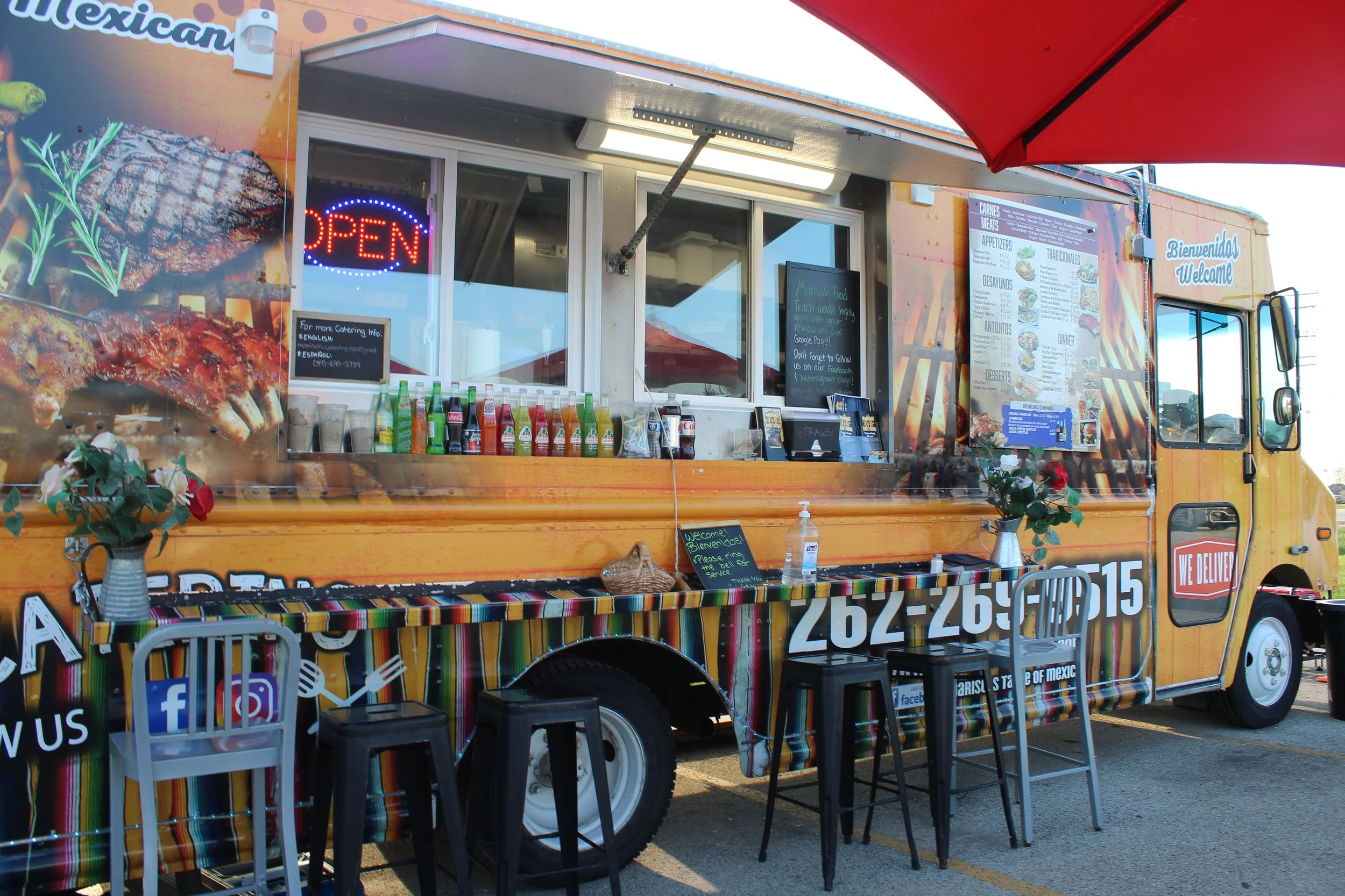 Mexican food food truck with colorful striped tablecloth, bottles of soda, flowers in vases, and a menu board on the side, in a parking lot.