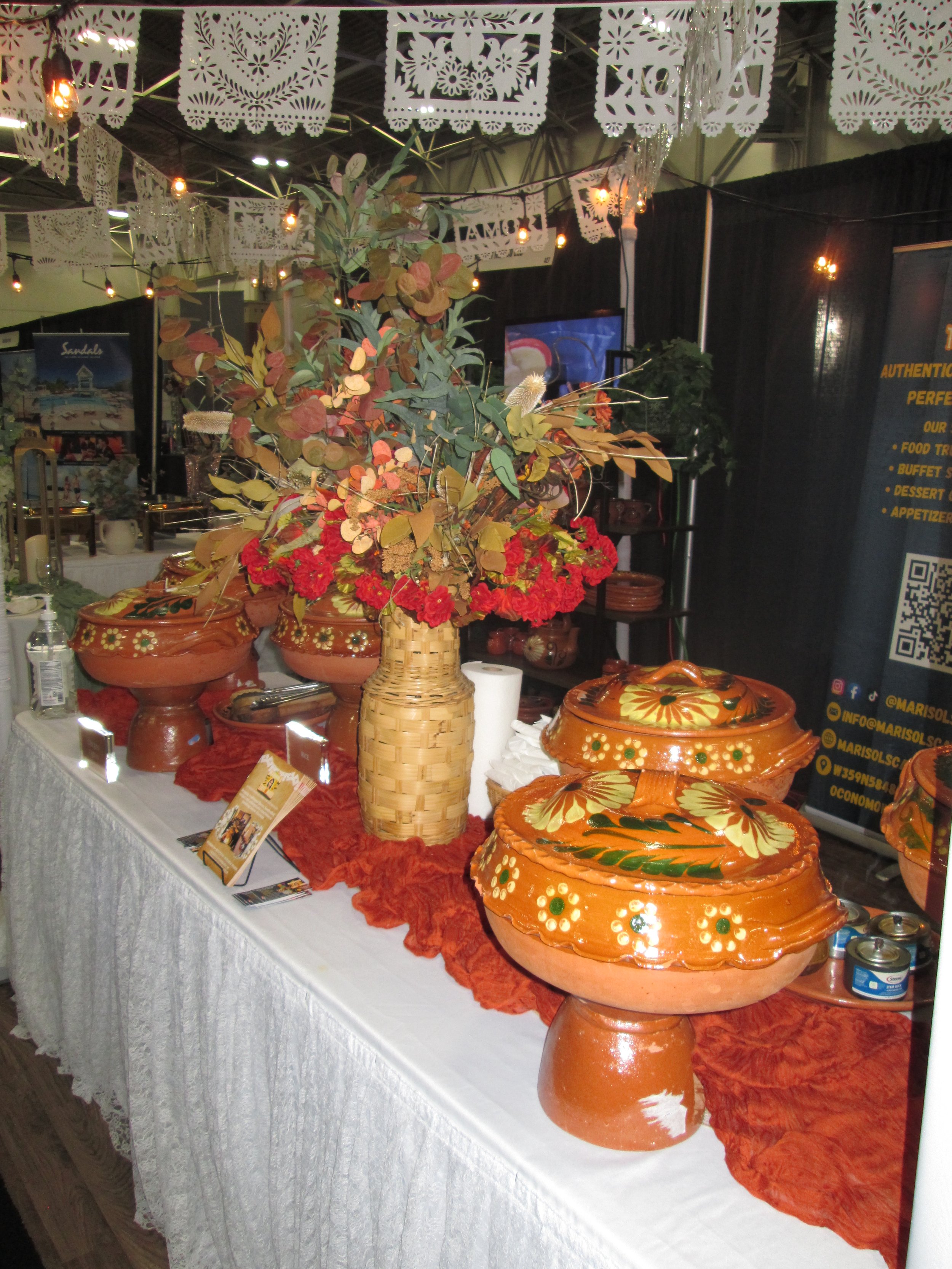 A display table with colorful ceramic pottery, a large woven basket, a bouquet of flowers and leaves in a vase, and informational pamphlets. The background features hanging paper decorations and banners at an indoor event, with some lights and signage visible.