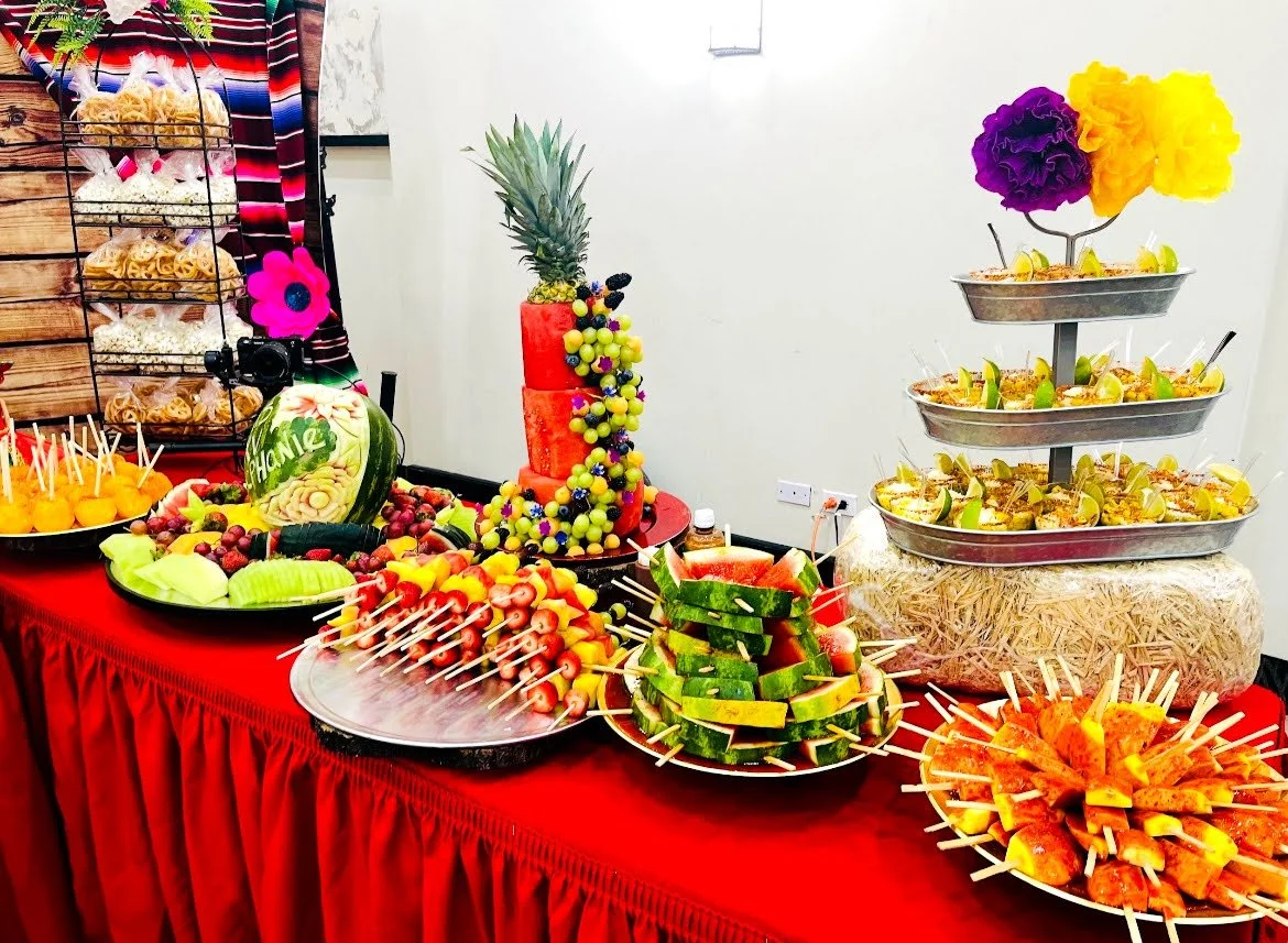 An assortment of colorful fruits and desserts arranged on a table with a red tablecloth, including a carved watermelon, a pineapple, a tiered tray with various mini desserts topped with flowers, and skewers of fruits like strawberries, melons, and watermelon.