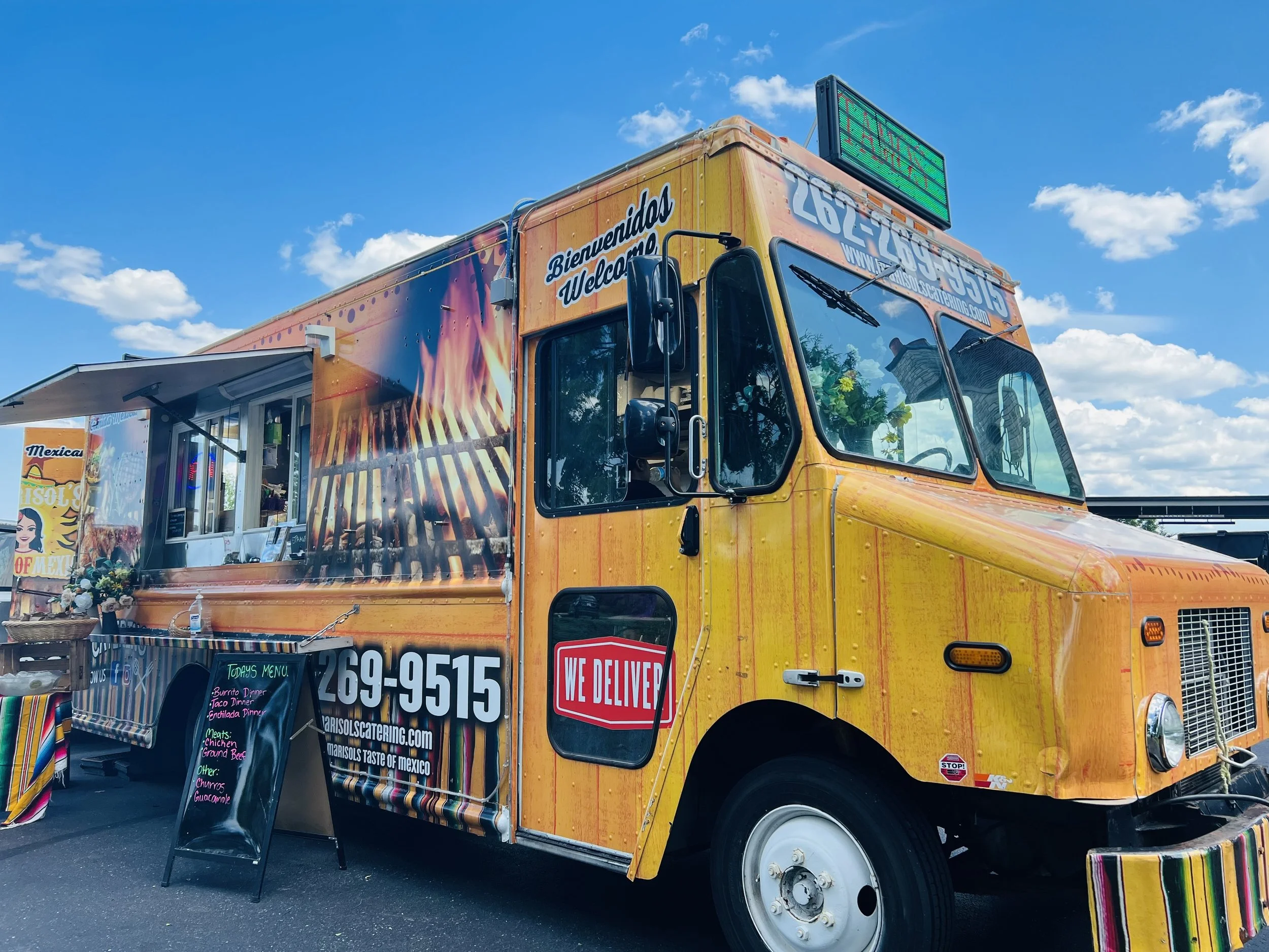 Food truck with colorful taco branding, menu board, and open serving window, parked outdoors under a partly cloudy sky.