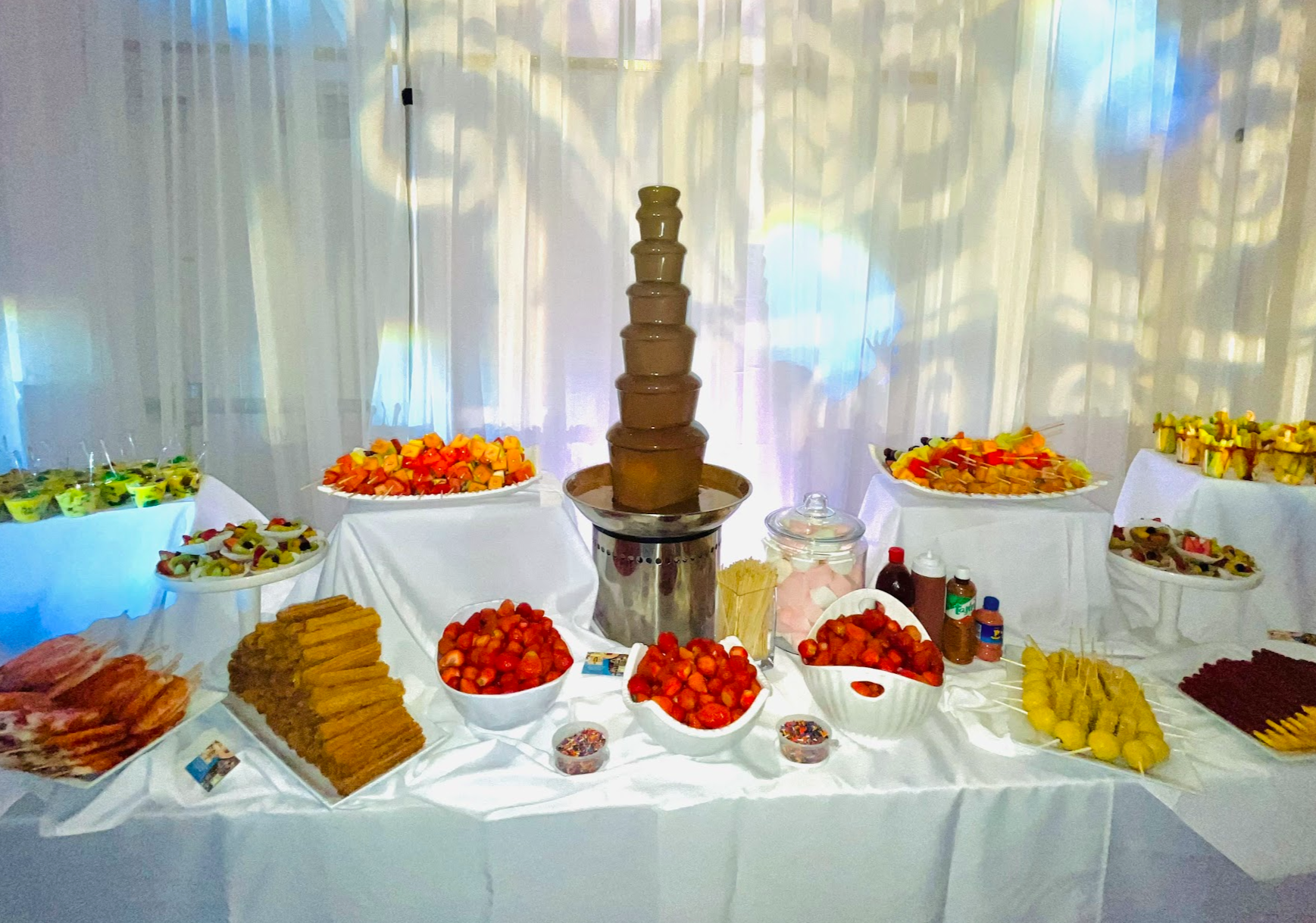 A dessert table with a chocolate fountain surrounded by bowls of strawberries, grapes, and assorted fruits, with plates of marshmallows, cookies, and skewered fruits, set against a white curtained backdrop.