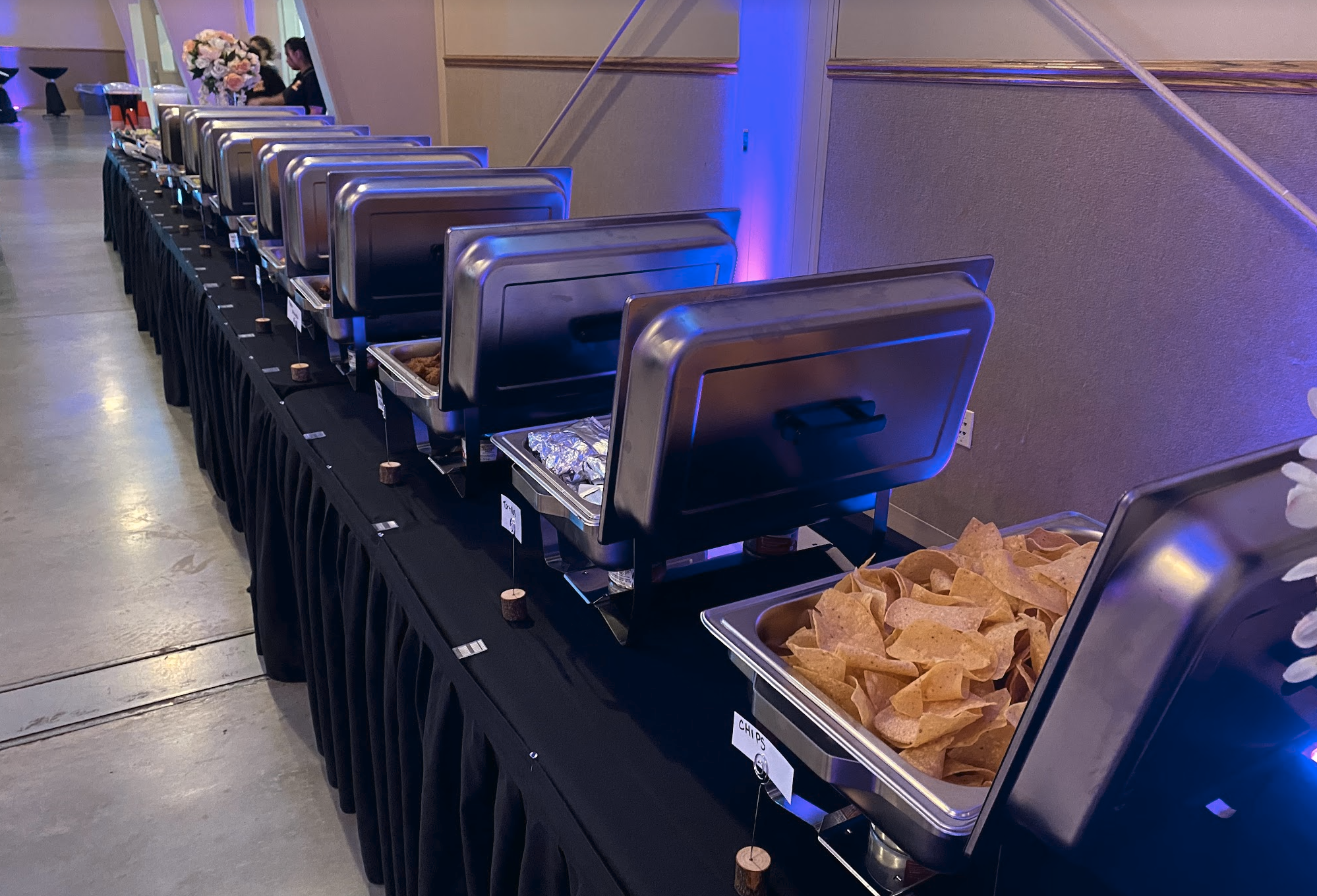 Buffet table with trays of chips, some covered with silver lids, in a dimly lit room with purple lighting.