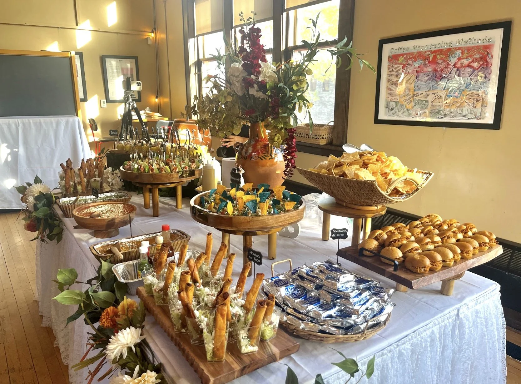 A buffet table with various appetizers and snacks, decorated with flowers and a large vase, in a well-lit room with framed wall art and large windows.