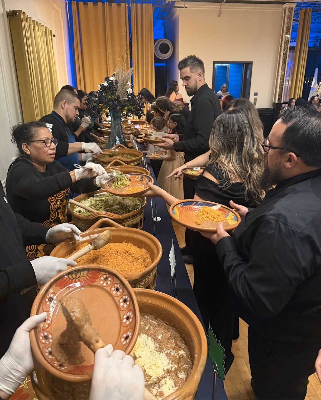 People lined up at a buffet serving traditional Mexican dishes like tamales, rice, and guacamole in a festive setting.