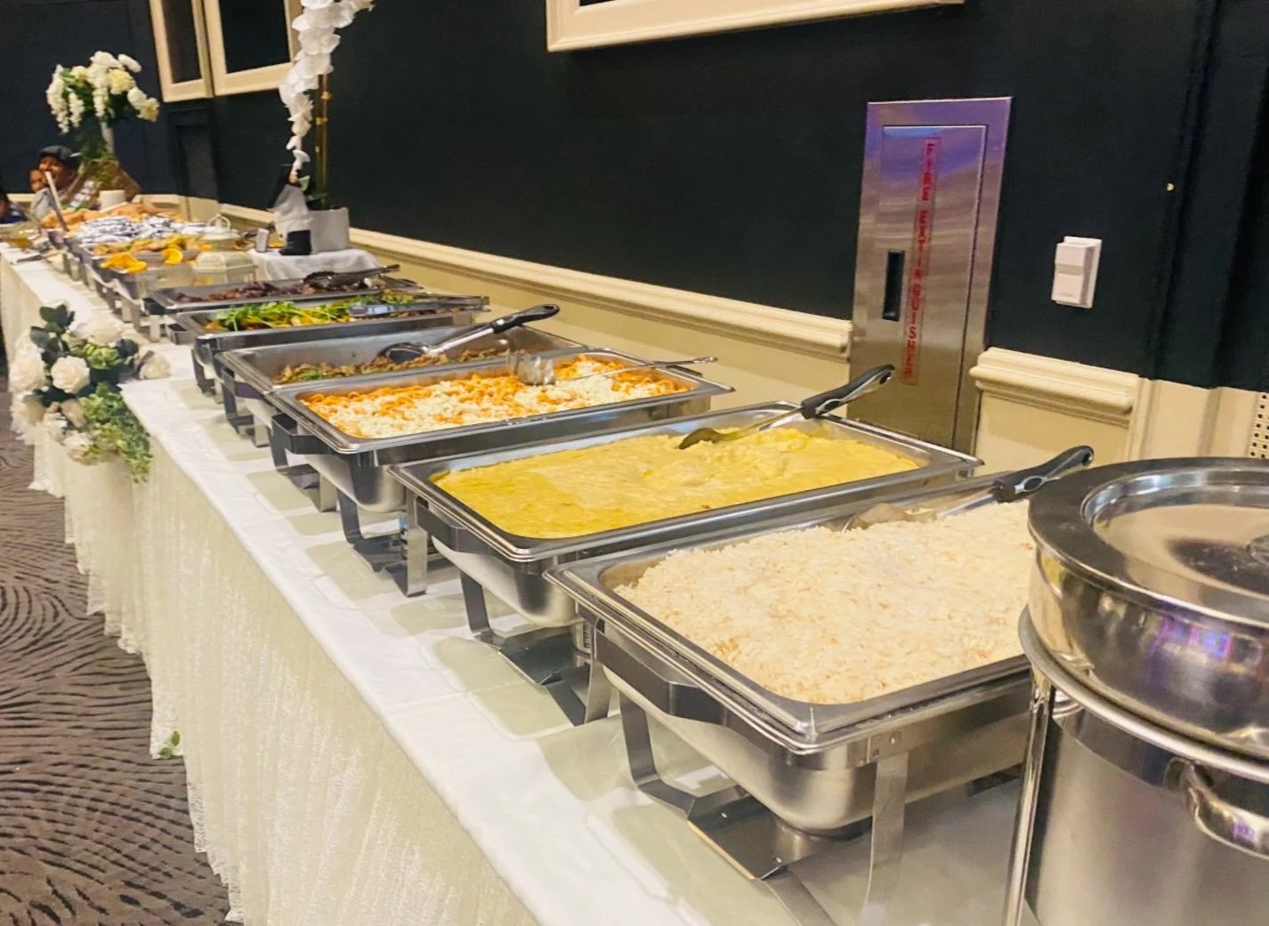 Buffet table with various trays of food including rice, pasta, vegetables, and curry, decorated with flowers in a banquet hall.