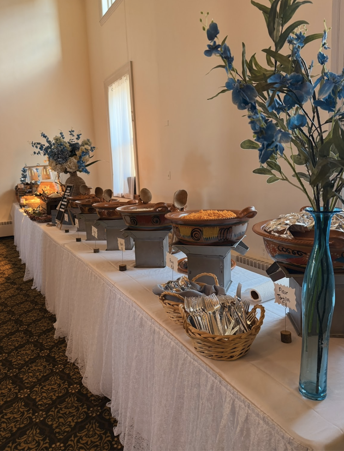 A buffet table with various dishes, bowls, and utensils, decorated with floral arrangements in vases, set in a room with beige walls and large windows.