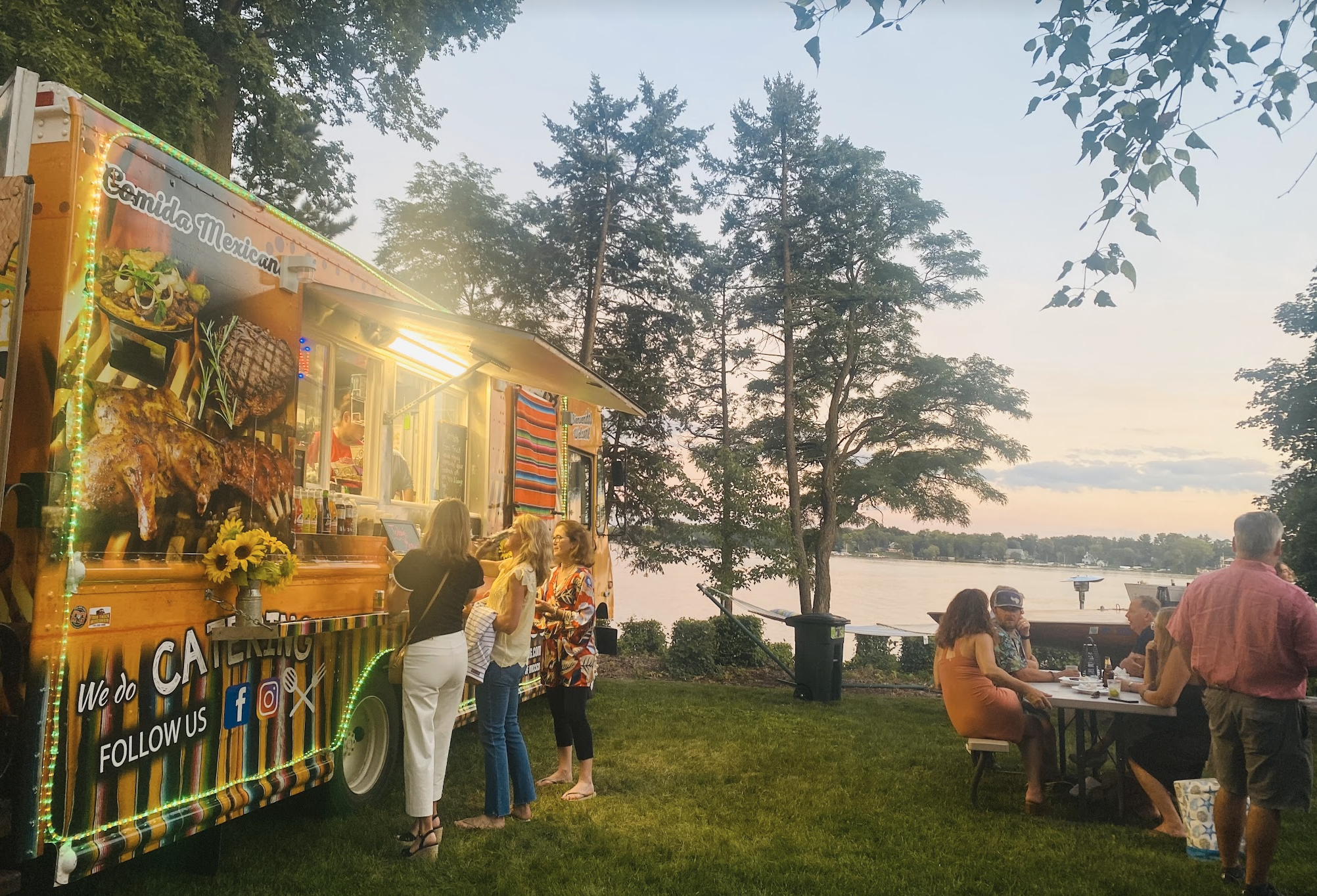 A Mexican food food truck serving grilled meats and tacos, with people ordering and enjoying food outdoors by a lake at sunset.