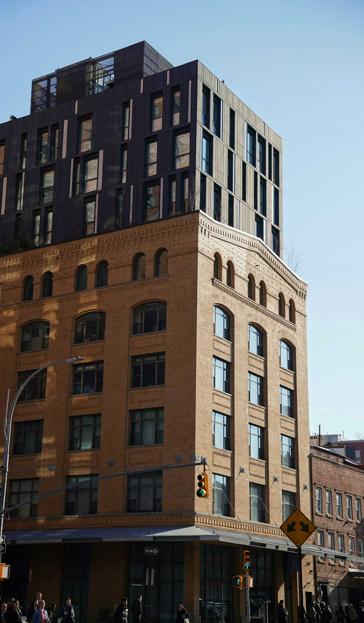 City street scene with a multi-story building featuring brick and modern black panels, people walking on the sidewalk, and traffic lights in the foreground.