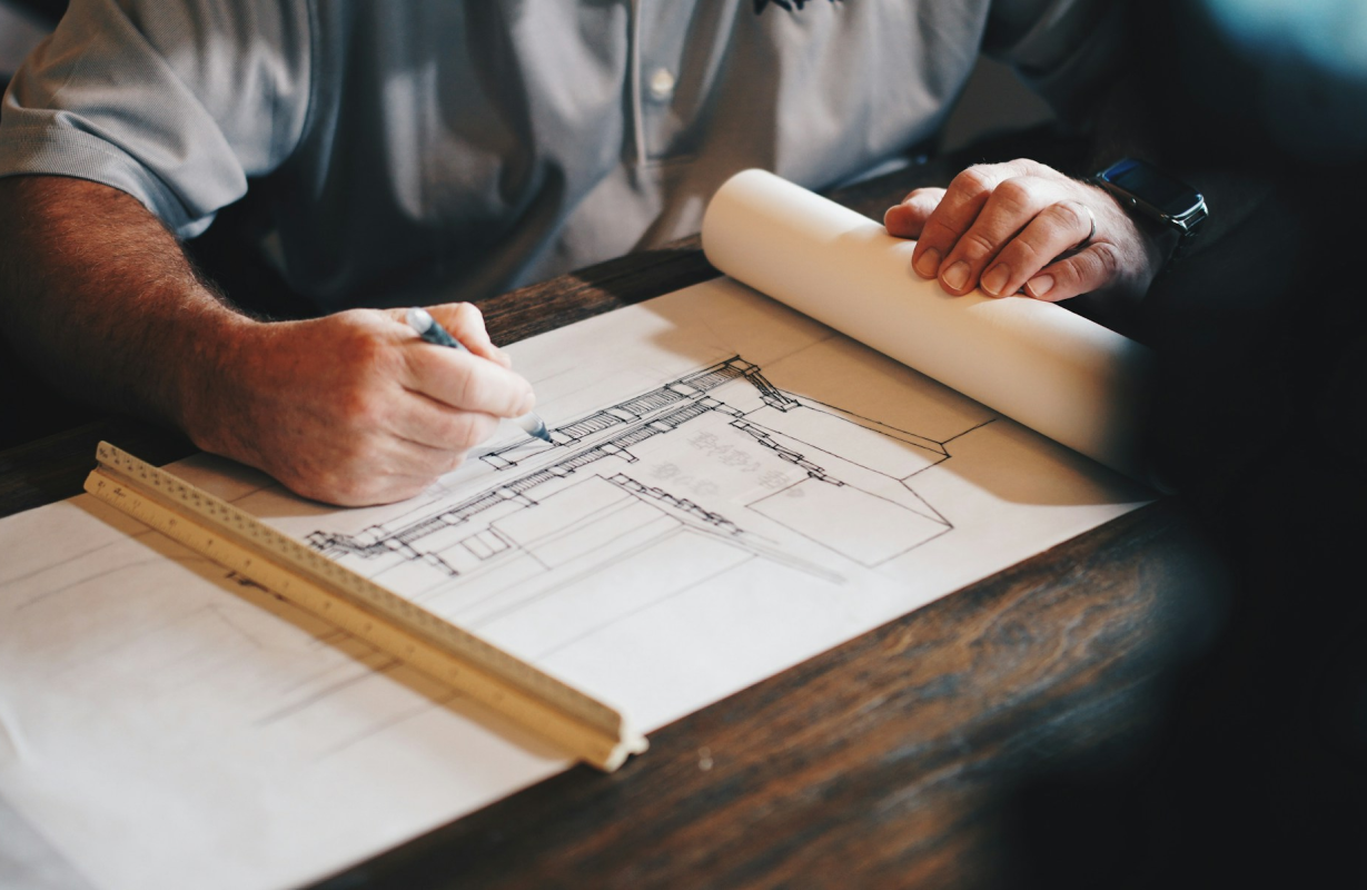 A person in a gray shirt is drawing architectural plans on paper with a black pen, using a wooden ruler on a dark wooden table.