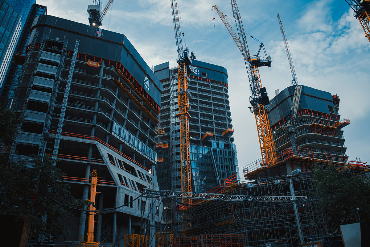 Under construction high-rise buildings with cranes, scaffolding, glass windows, and steel framework, set against a partly cloudy sky.
