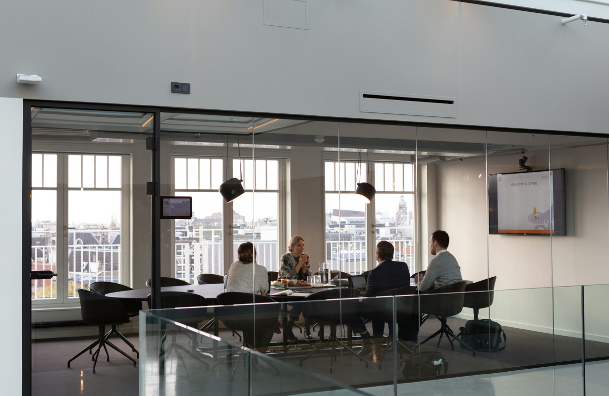 Business meeting inside a modern conference room with four people sitting around a table, viewed through glass walls, with city buildings visible outside.