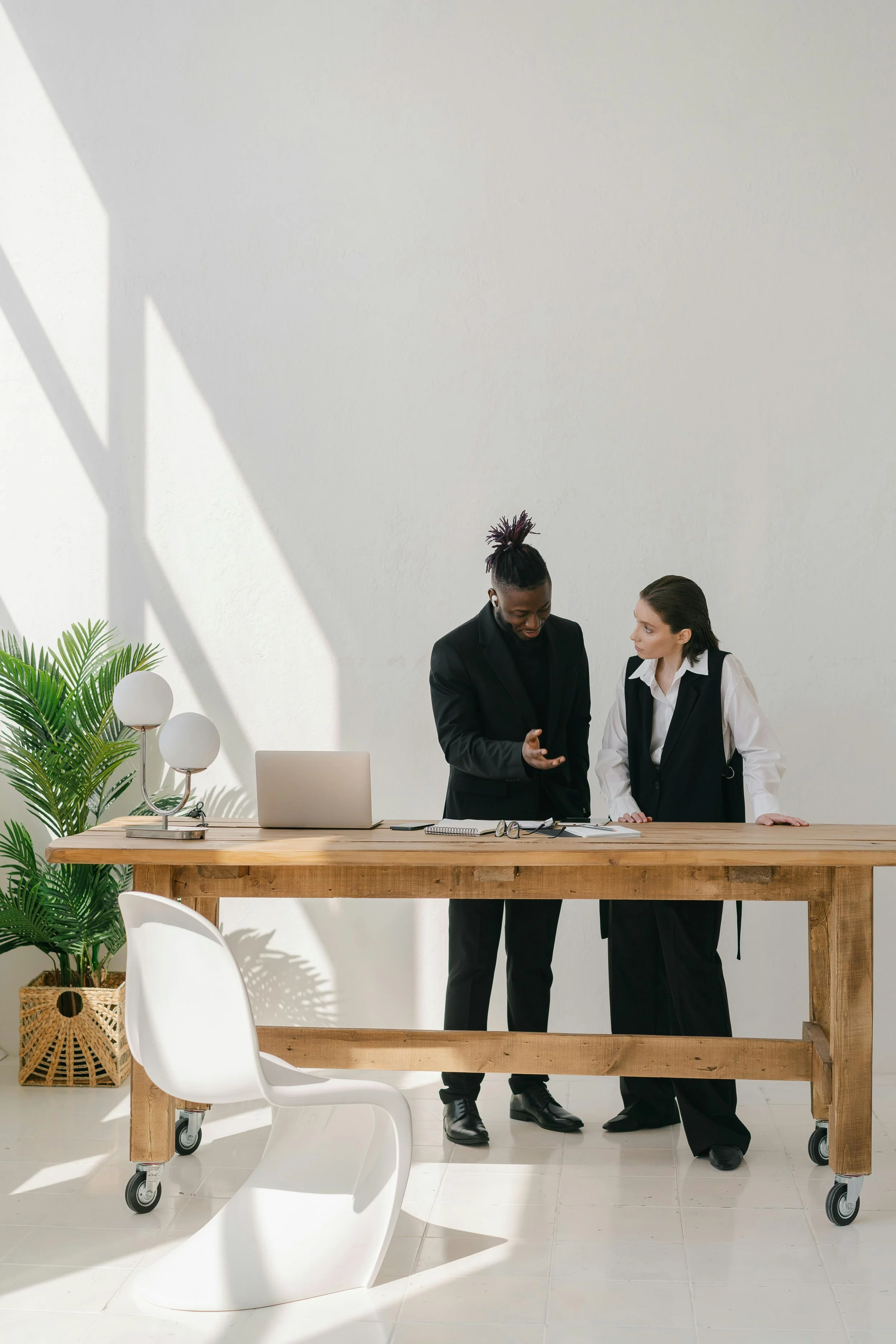 Two professionals, one African American and one Caucasian, are standing behind a wooden table in a bright, modern office space. They are engaged in a discussion, with the man gesturing with his hand. The table has a laptop, glasses, and some papers. There is a white aesthetic chair in the foreground, a large green plant on the left, and a white wall with a shadow pattern in the background.
