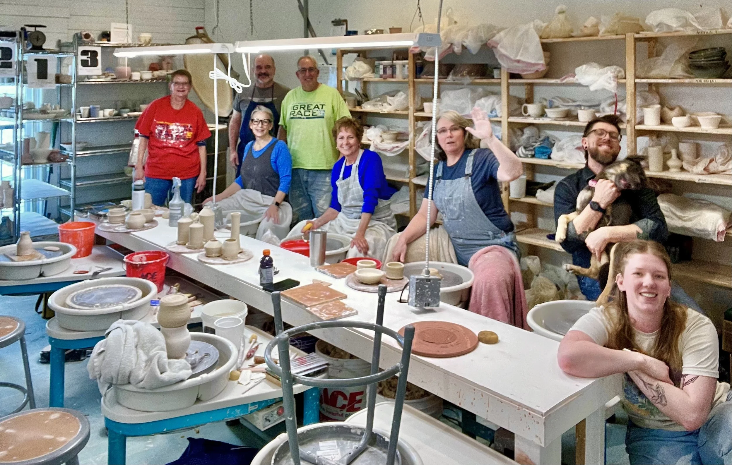 A group of eight people and one dog pose in a pottery studio with shelves of unglazed ceramics behind them and tables with pottery supplies in front.